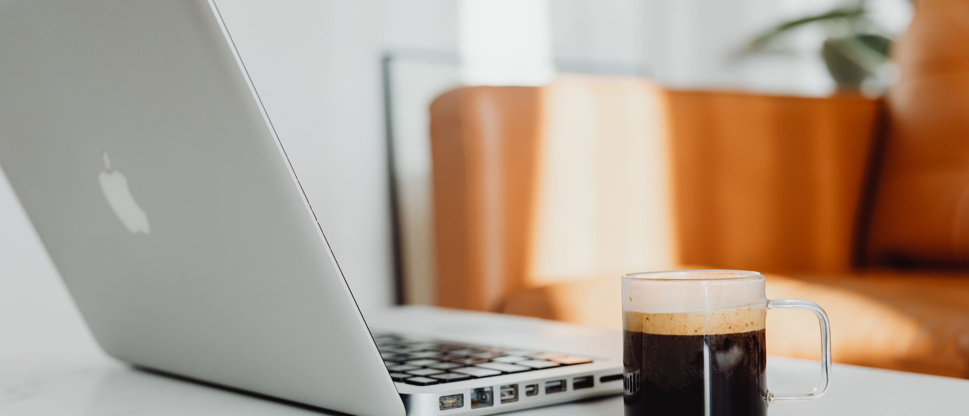 Close-up of a silver Apple MacBook on a white desk, with a clear glass mug filled with black coffee and topped with foam in the foreground. In the background, an orange armchair and a potted plant are visible.