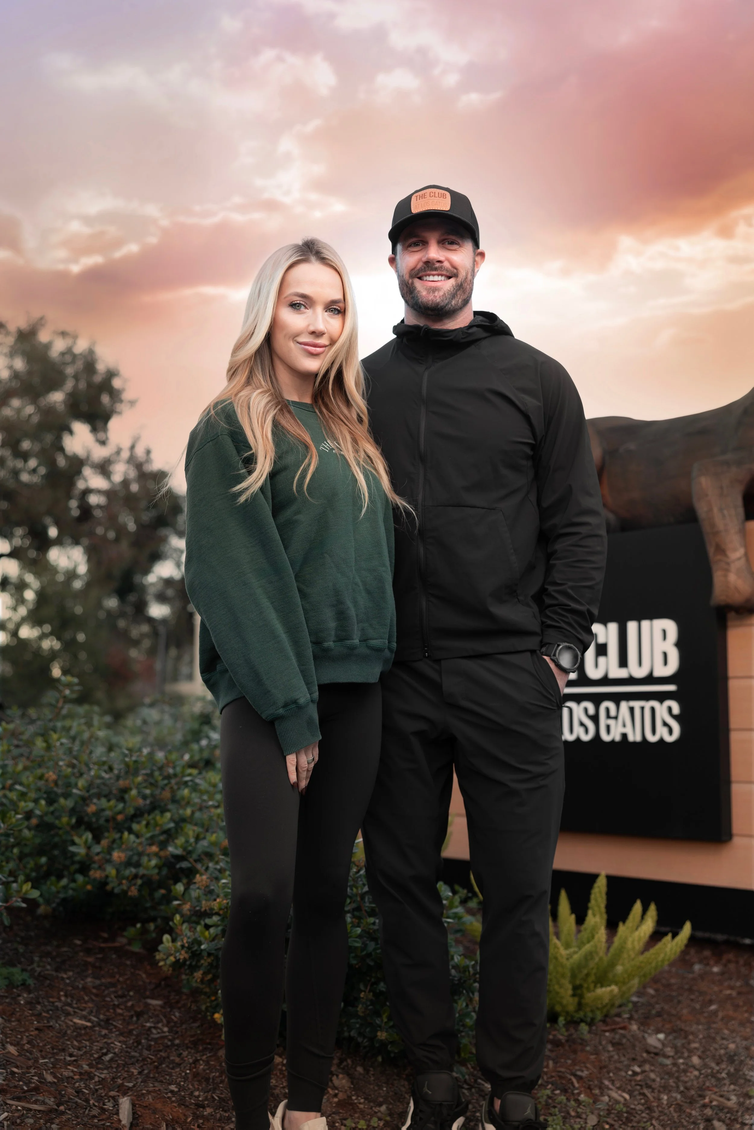 Two local owners that are partners outdoors at sunset, smiling at the camera - There are trees, bushes, and a sign that reads 'THE CLUB' in the background.
