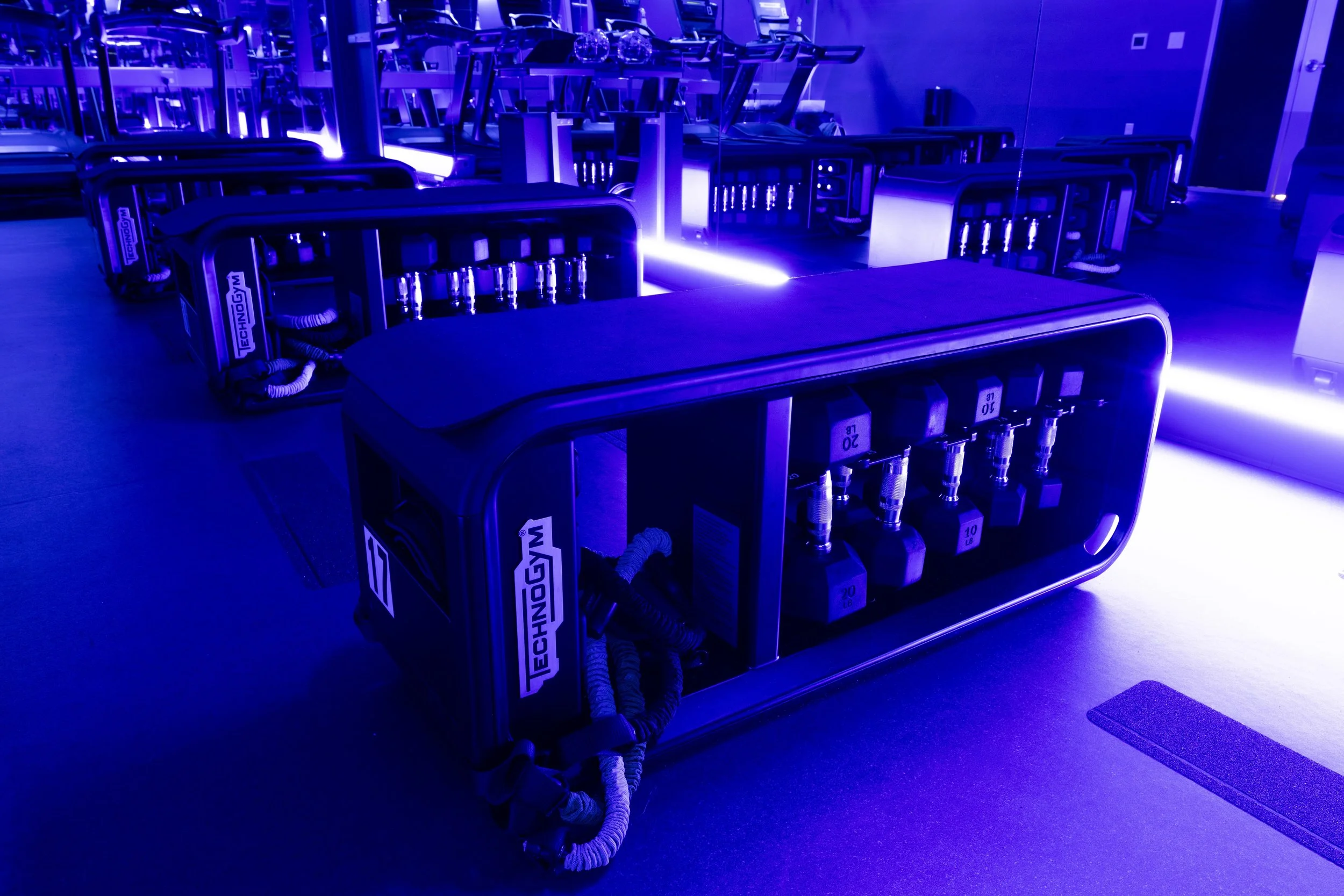 Equipment for a gym or fitness center, illuminated with blue lighting, including rows of exercise machines and weights.