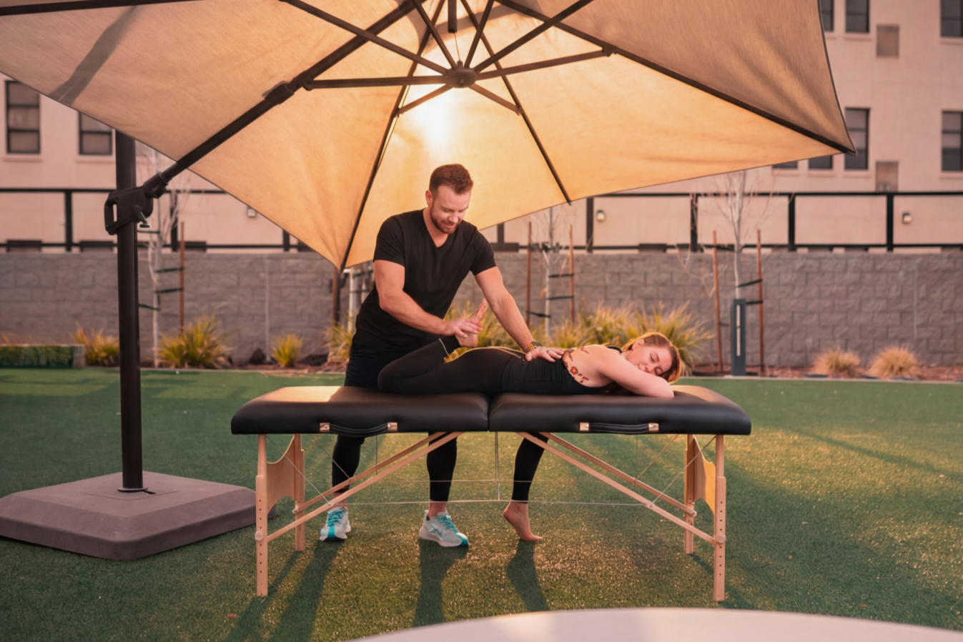 A man giving a massage to a woman lying face down on a massage table outdoors under a large umbrella.