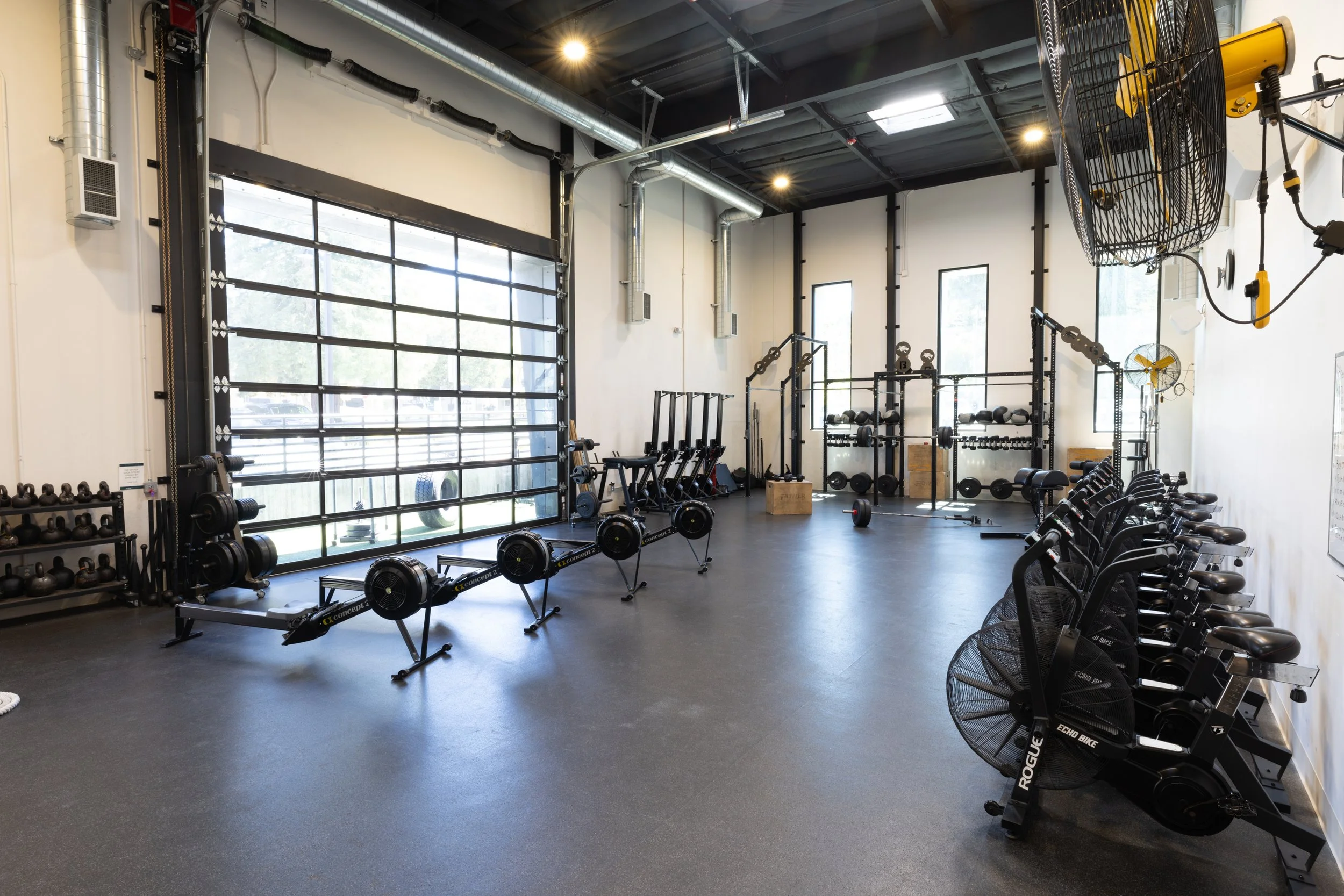 An empty modern gym with black rubber flooring, a large garage-style glass door, and various workout equipment including kettlebells, rowing machines, weight racks with dumbbells, and fans.