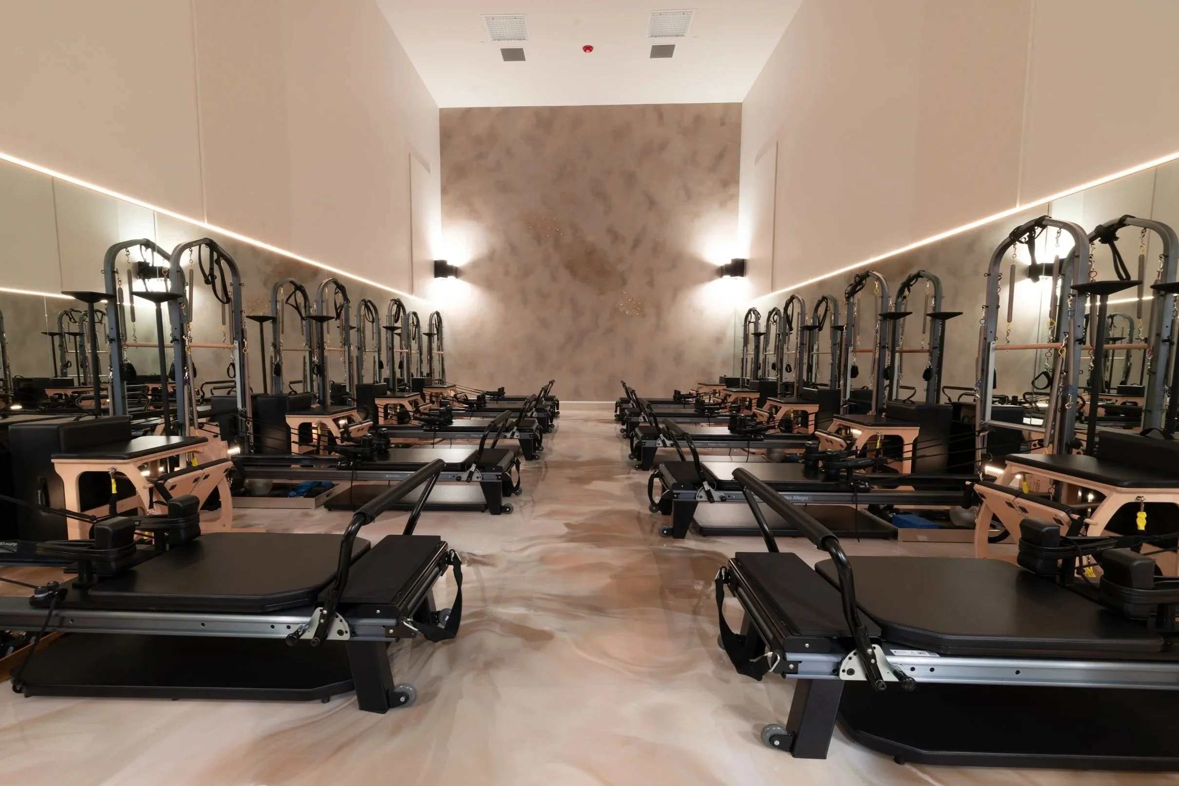 A Pilates or fitness studio with multiple reformer machines lined up in rows against a stone-textured wall, illuminated by wall-mounted lights and LED strip lighting along the ceiling edges.