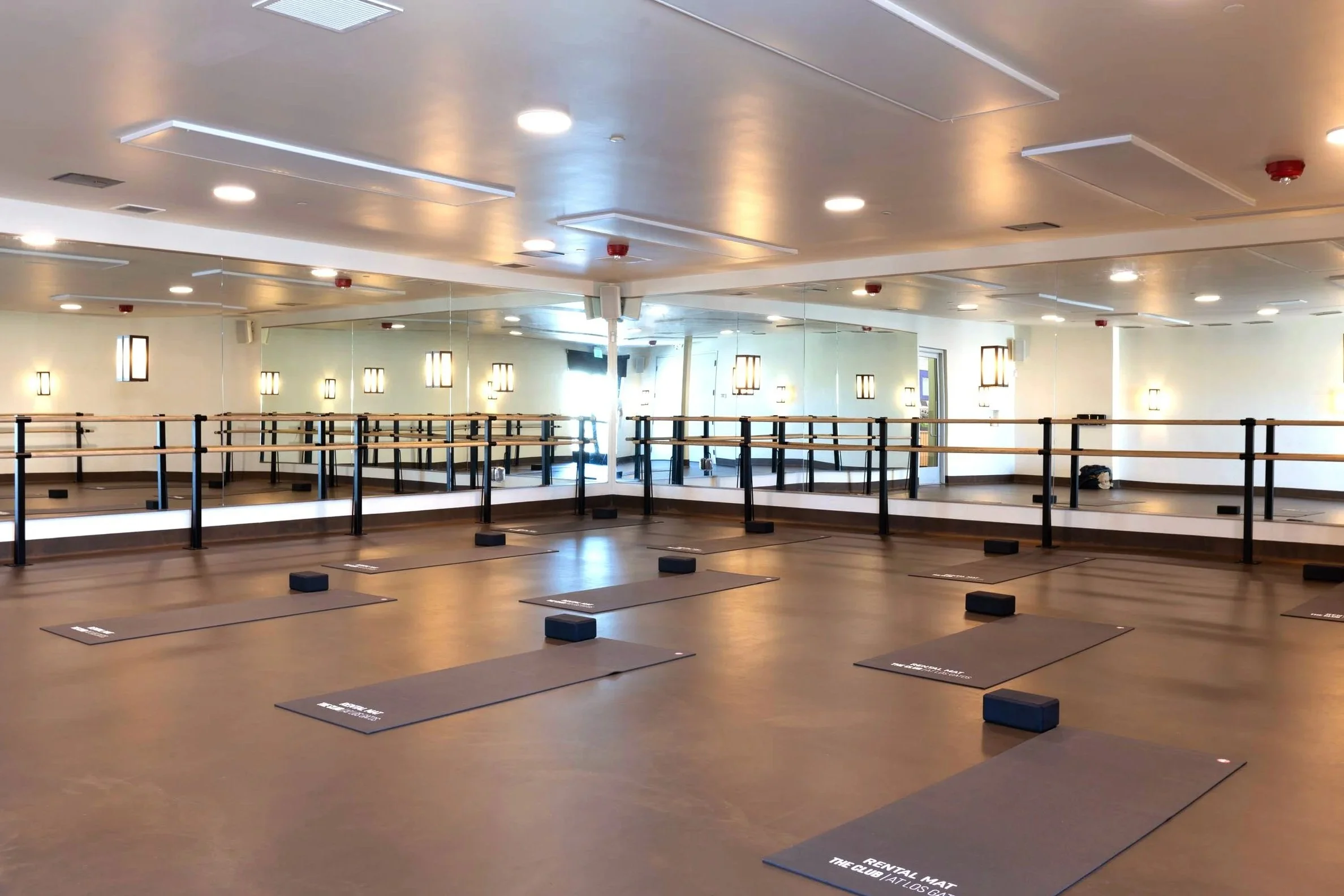 Interior of a dance or yoga studio with mirrors on the wall, wooden flooring, black mats with white text, foam blocks, and ballet barres, illuminated by ceiling lights and wall sconces.