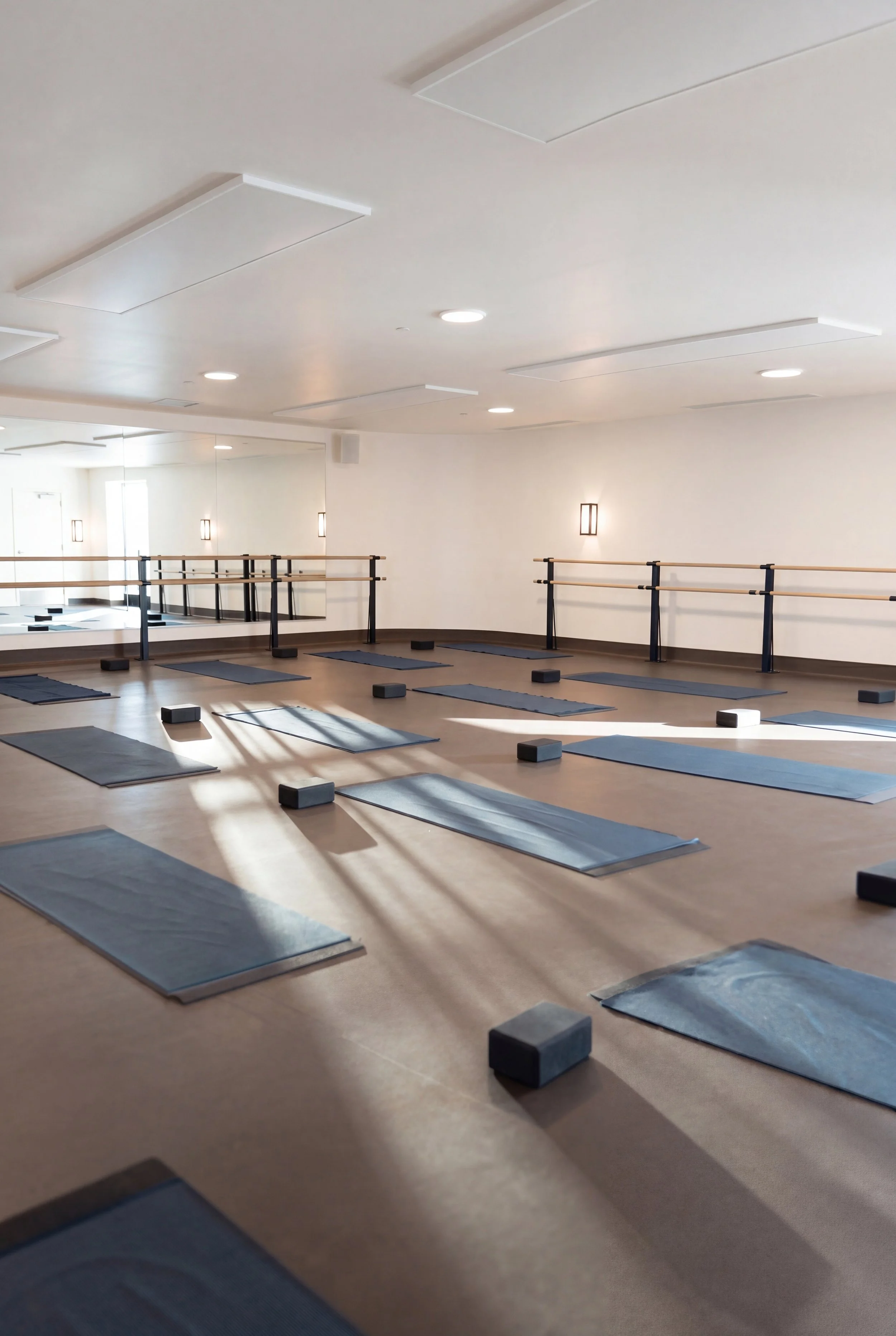 A spacious yoga studio with yoga mats and blocks arranged on the floor, mirrors on the wall, ballet barres along the wall, and soft lighting.