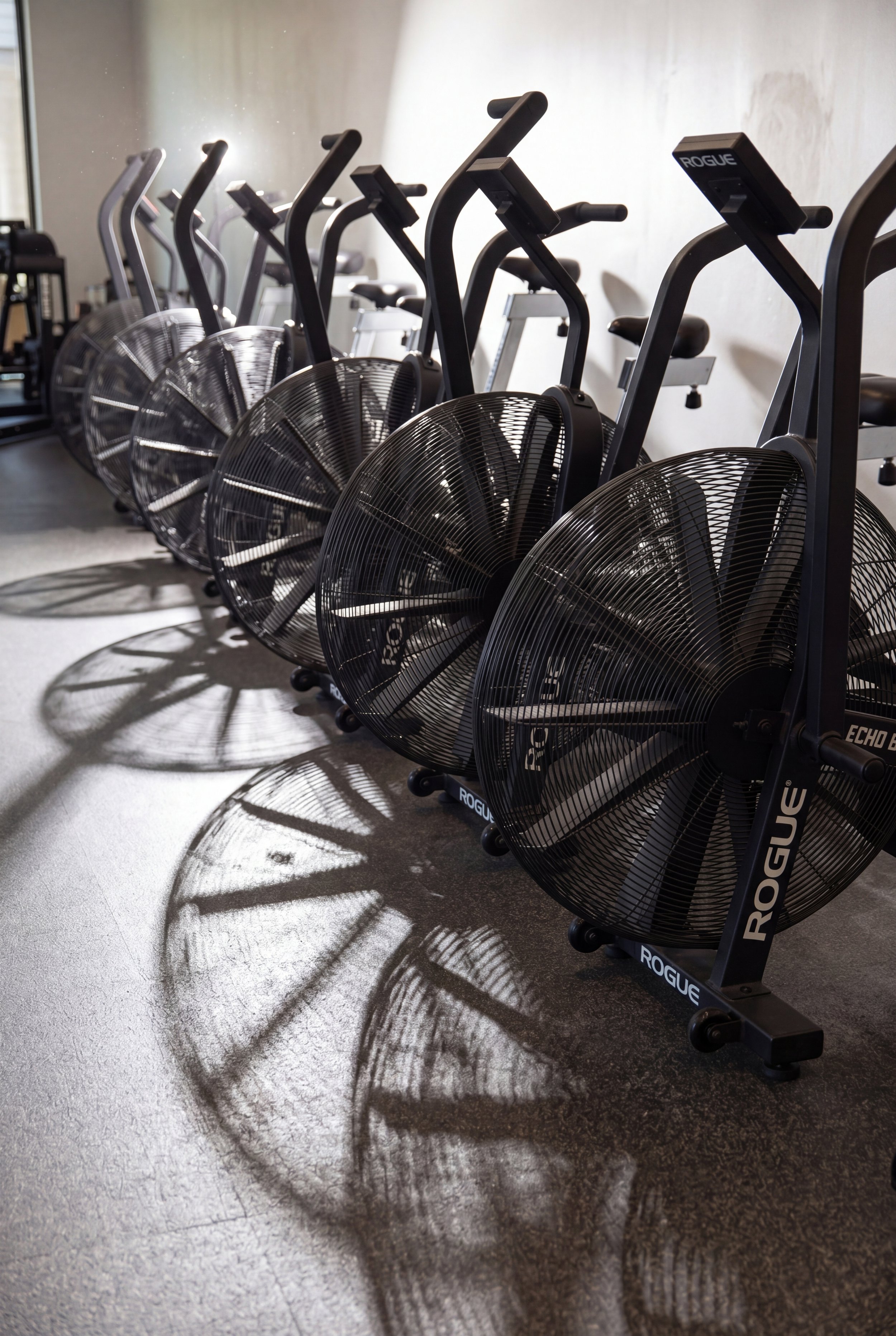 Row of elliptical exercise bikes with large front fans casting circular shadows on the floor, in a gym with a plain beige wall.