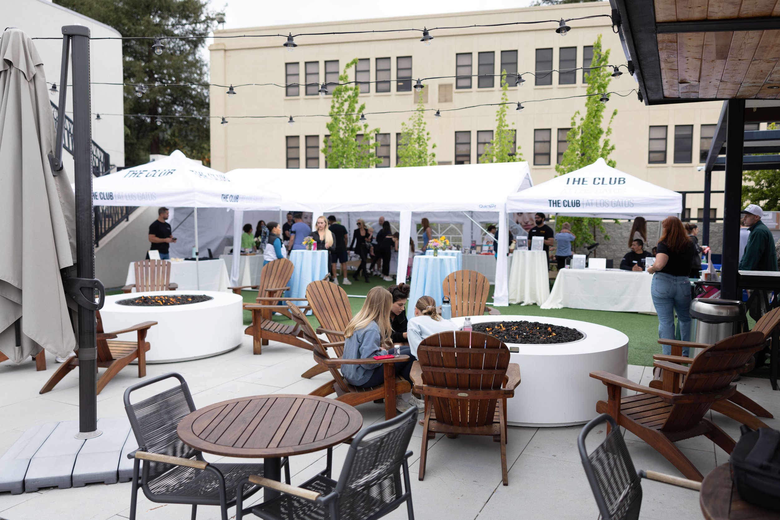 People gathered outdoors at a patio event with tents, tables, and string lights, surrounded by modern buildings and trees.