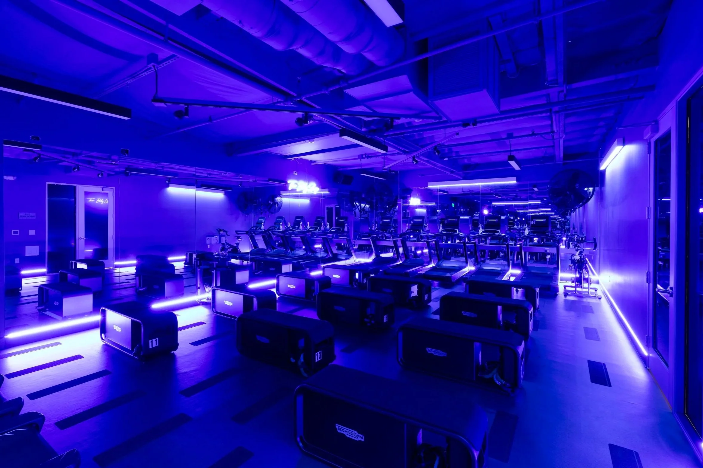 Indoor gym with workout stations illuminated by blue neon lighting, featuring treadmills, bikes, and exercise benches.