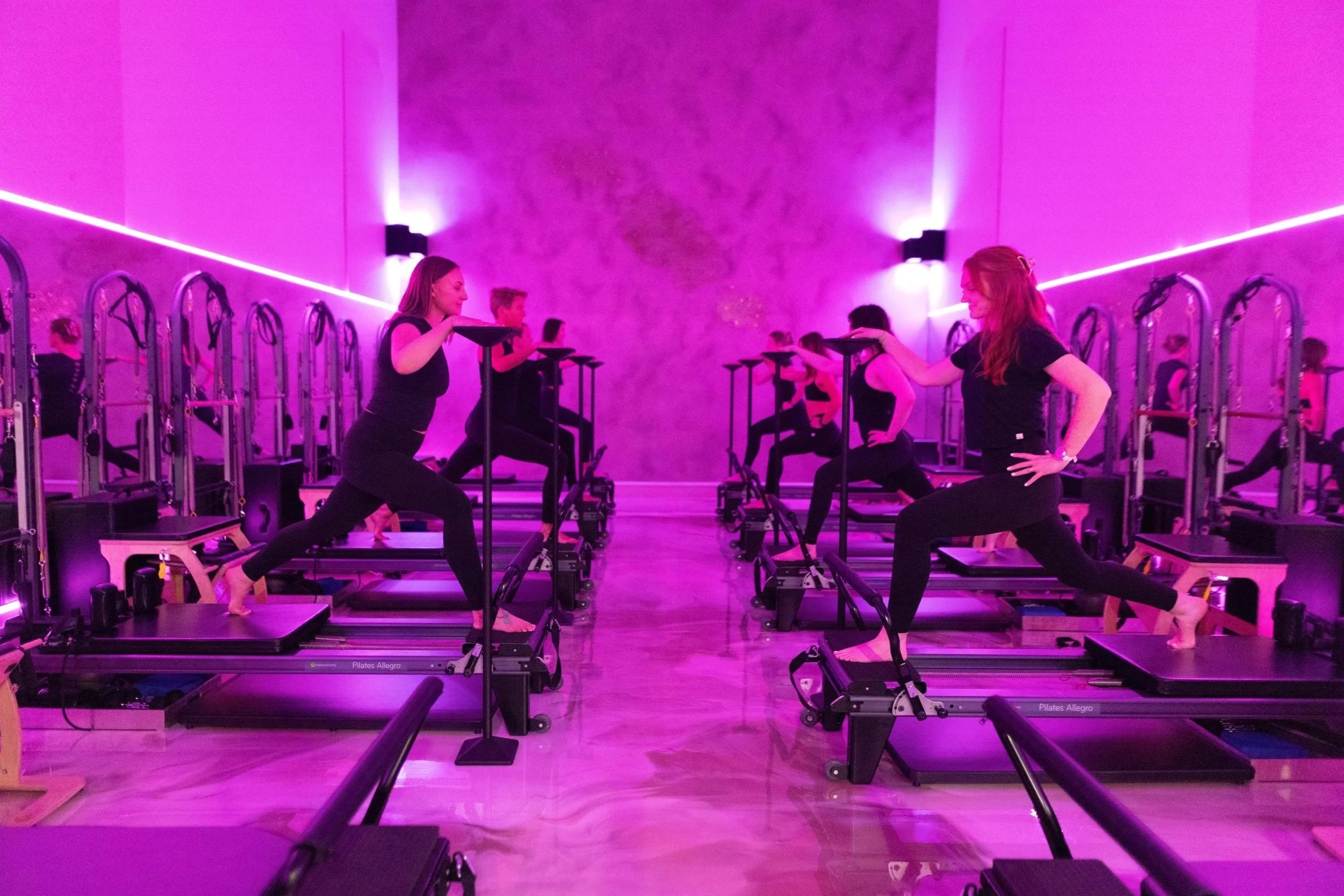 A group of people practicing Pilates in a dimly lit studio with purple lighting, using reformer machines.