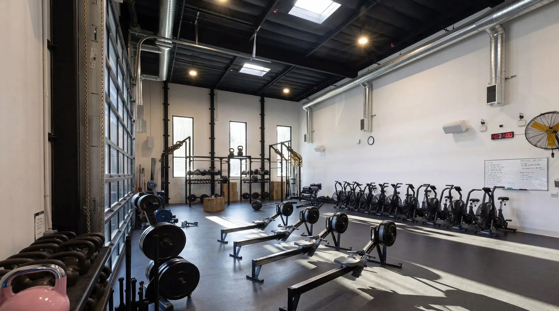 Interior view of a modern gym with black equipment, including rowing machines, stationary bikes, weight racks, kettlebells, and functional training equipment, illuminated by natural light from skylights and windows.