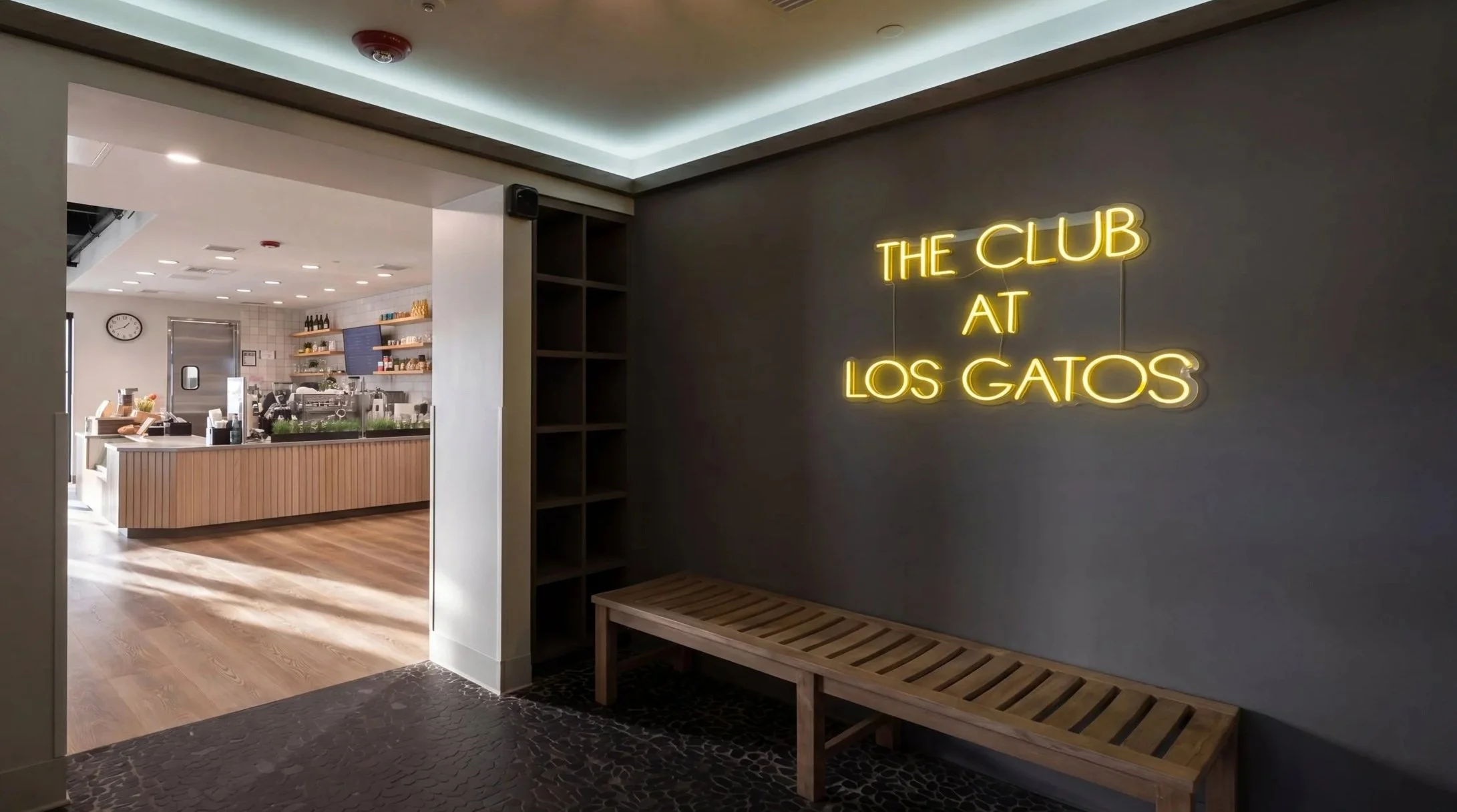 Interior view of a modern lounge or café with a neon sign that reads 'The Club at Los Gatos' on a gray wall, a wooden bench underneath, and a glimpse of the counter and seating area in the background.