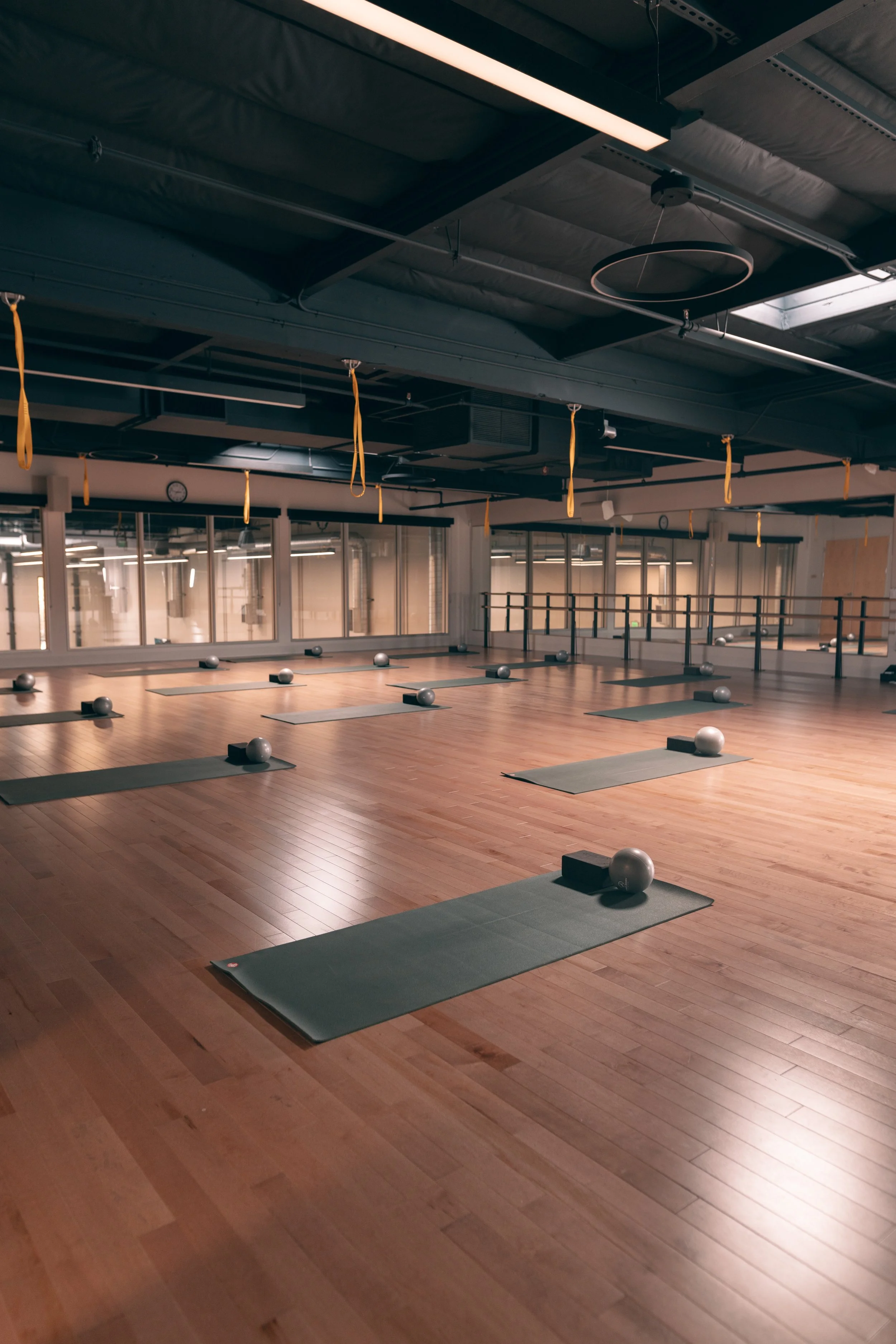 Yoga studio with wooden floors, yoga mats, silver exercise balls, and yellow resistance bands hanging from the ceiling. Large mezzanine window and mirrors are visible along one wall.