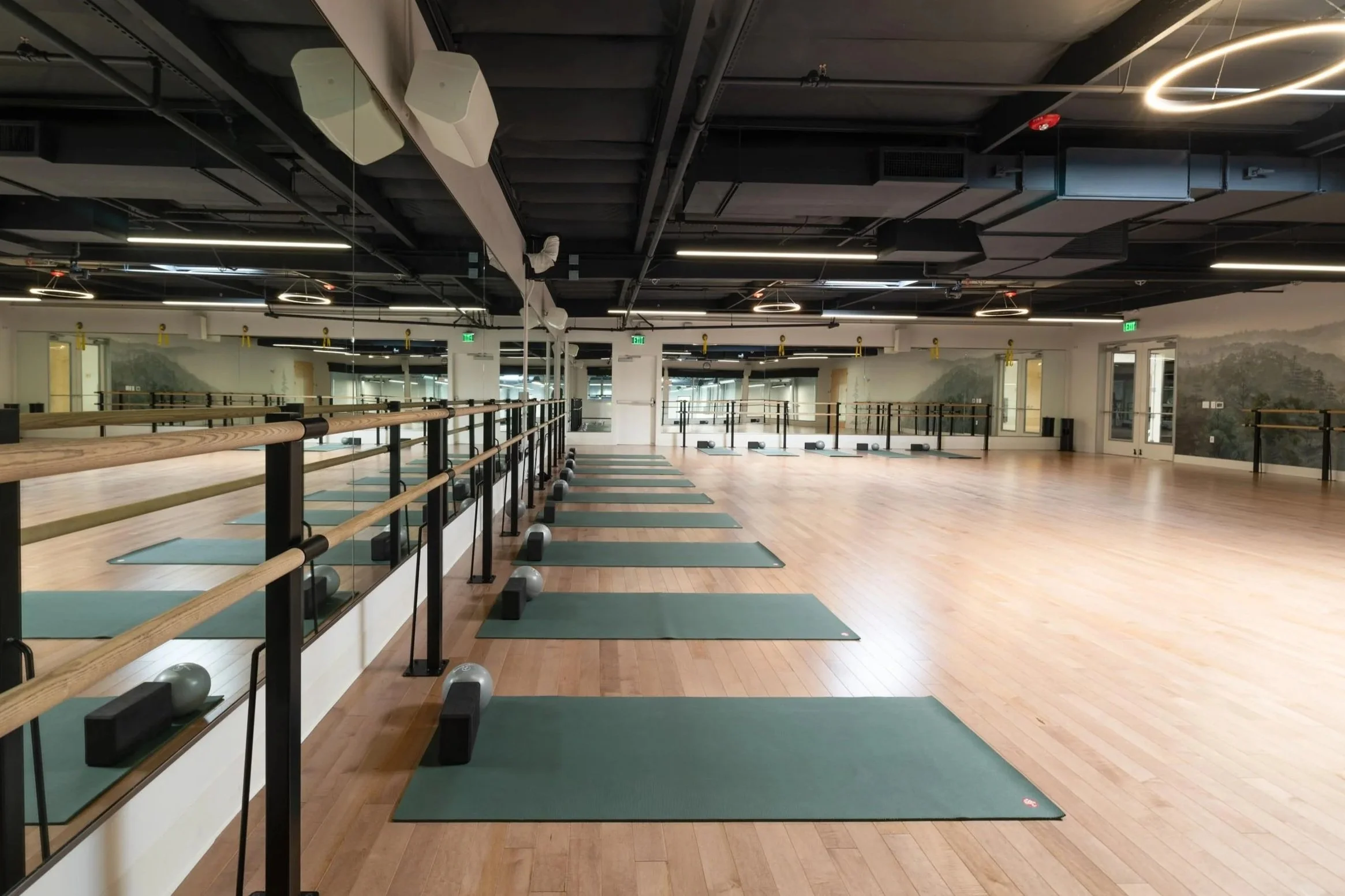 An empty dance or fitness studio with wooden floors, wall mirrors, and exercise mats on the floor. The ceiling has a modern black design with hanging lights and suspended speakers. Rooms with large windows and doors are visible along the walls.