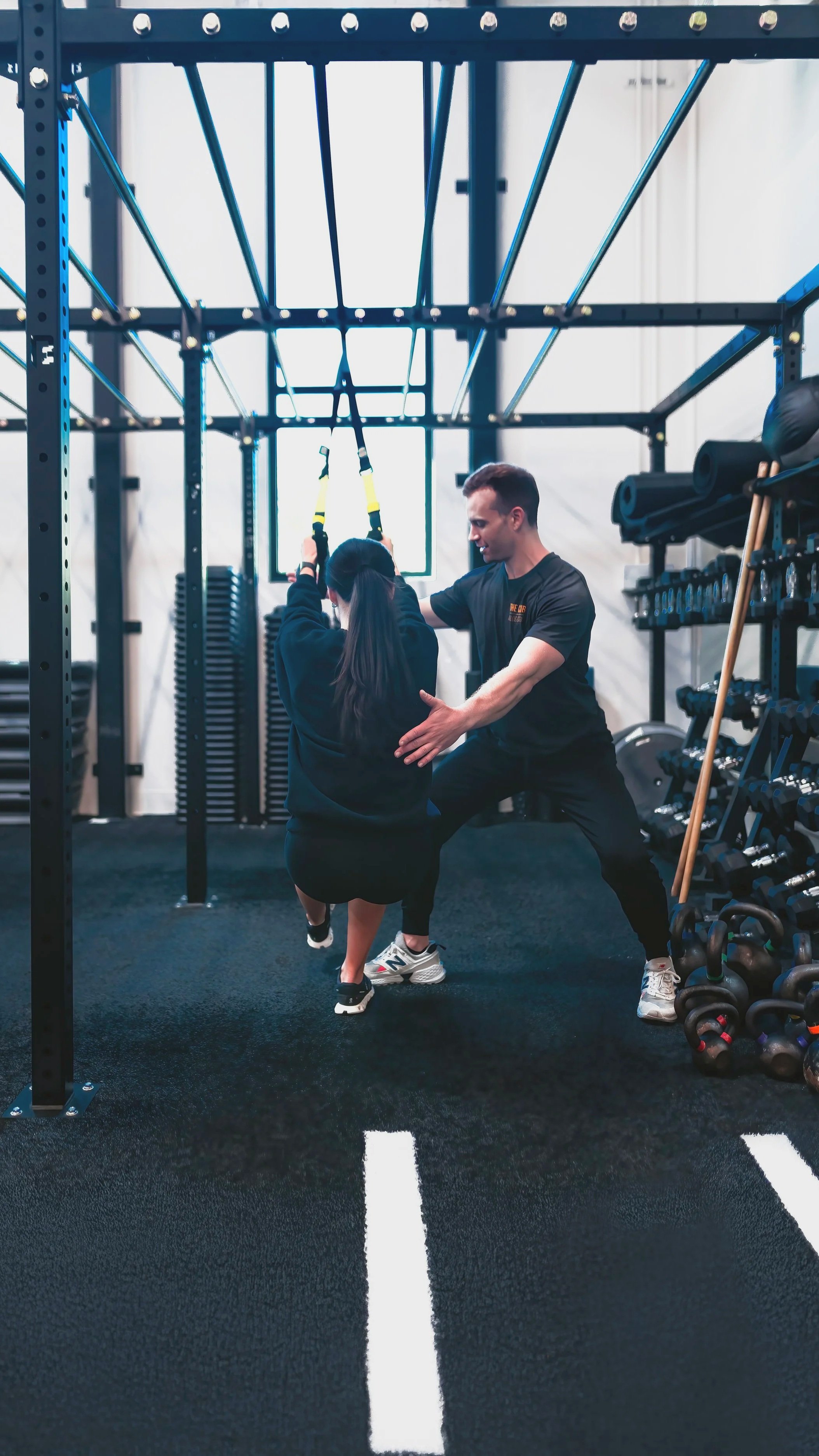 A personal trainer helping a woman perform a suspension exercise in a modern gym facility in a dedicated free-weight section with TRX straps, overhead rig structure, organized rack of kettlebells, dumbbells, range of functional training tools.