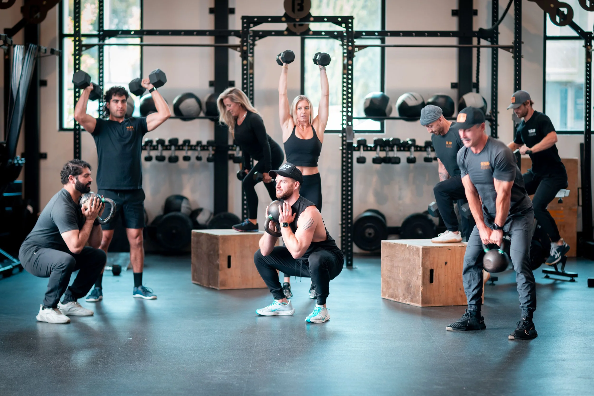 Group of people in a gym performing weightlifting exercises, some using kettlebells and dumbbells, with gym equipment and large windows in the background.