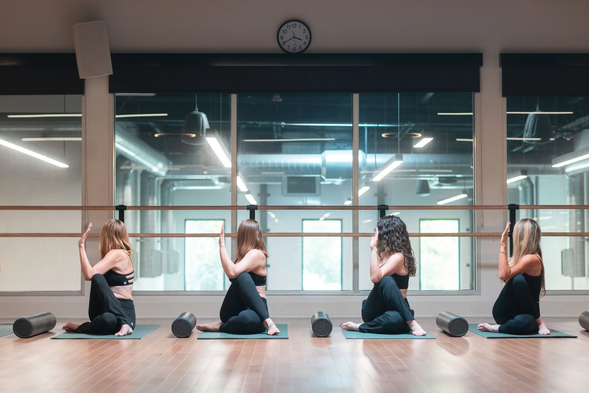 Women practicing yoga in a dance studio with large windows, mirrors, and wooden floors, seated on yoga mats with foam rollers nearby, raising their right hands.