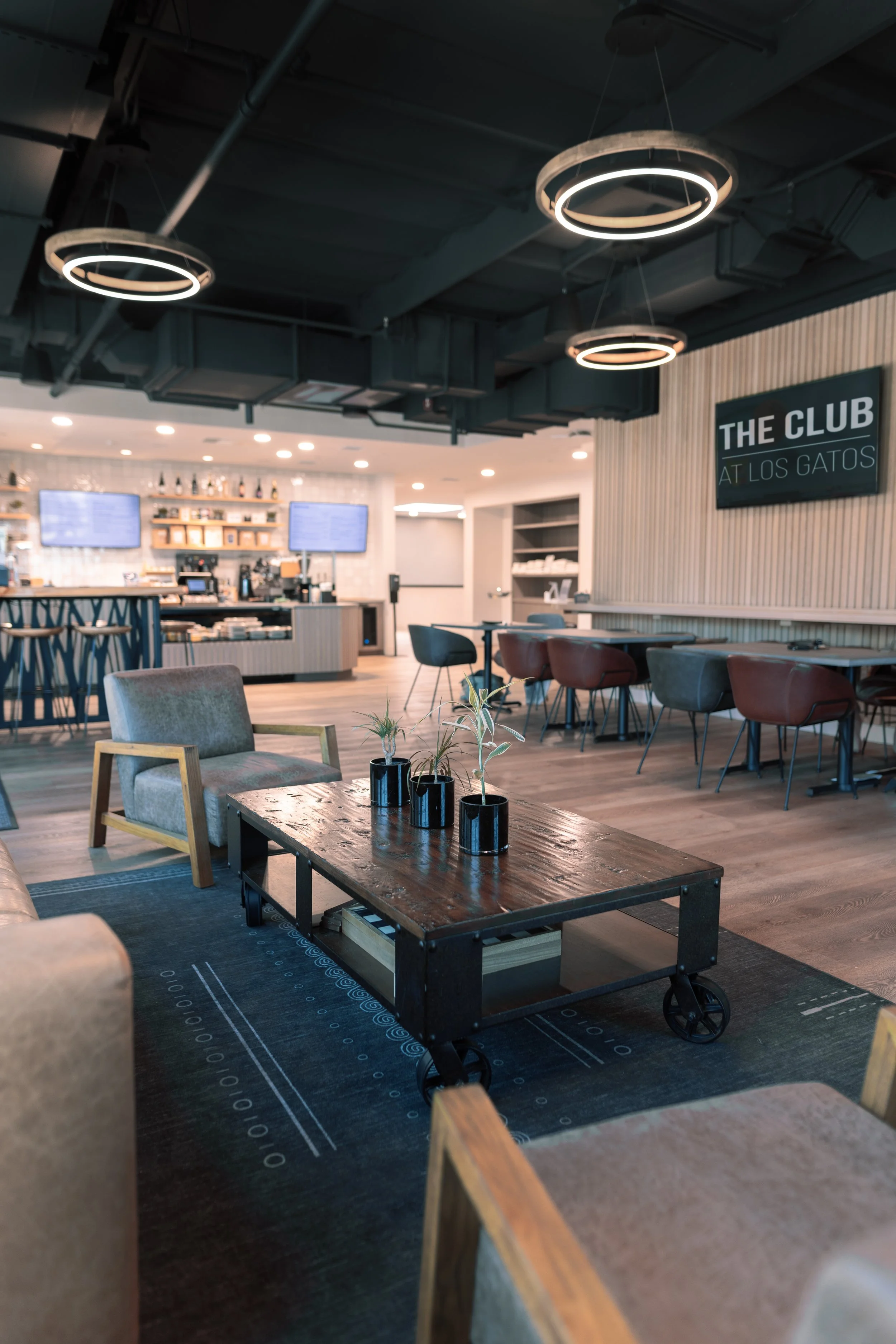 Modern cafe interior with wooden furniture, potted plants, and a black wagon-style coffee table. There are upholstered chairs and a rug on the floor, with a bar and menu screens in the background.