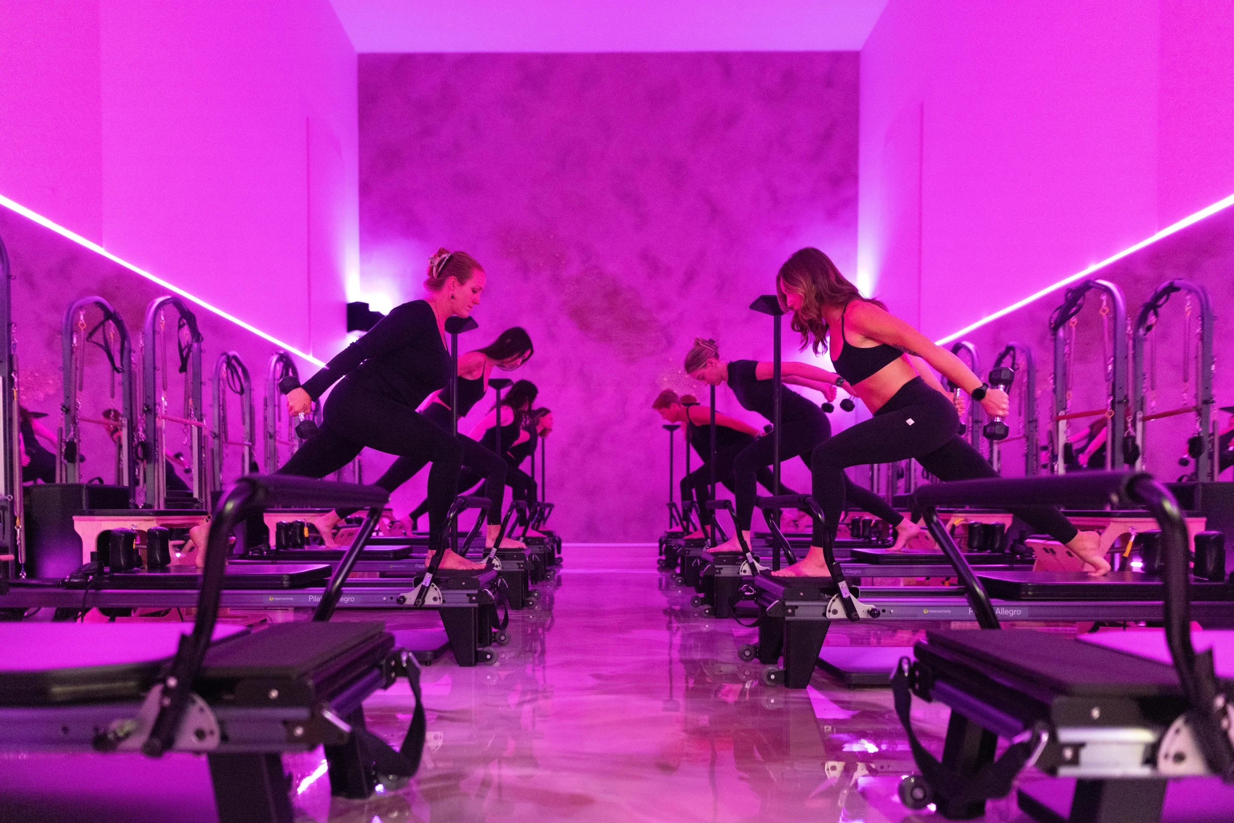 People attending a fitness class in a room with pink and purple lighting, using reformer Pilates machines.
