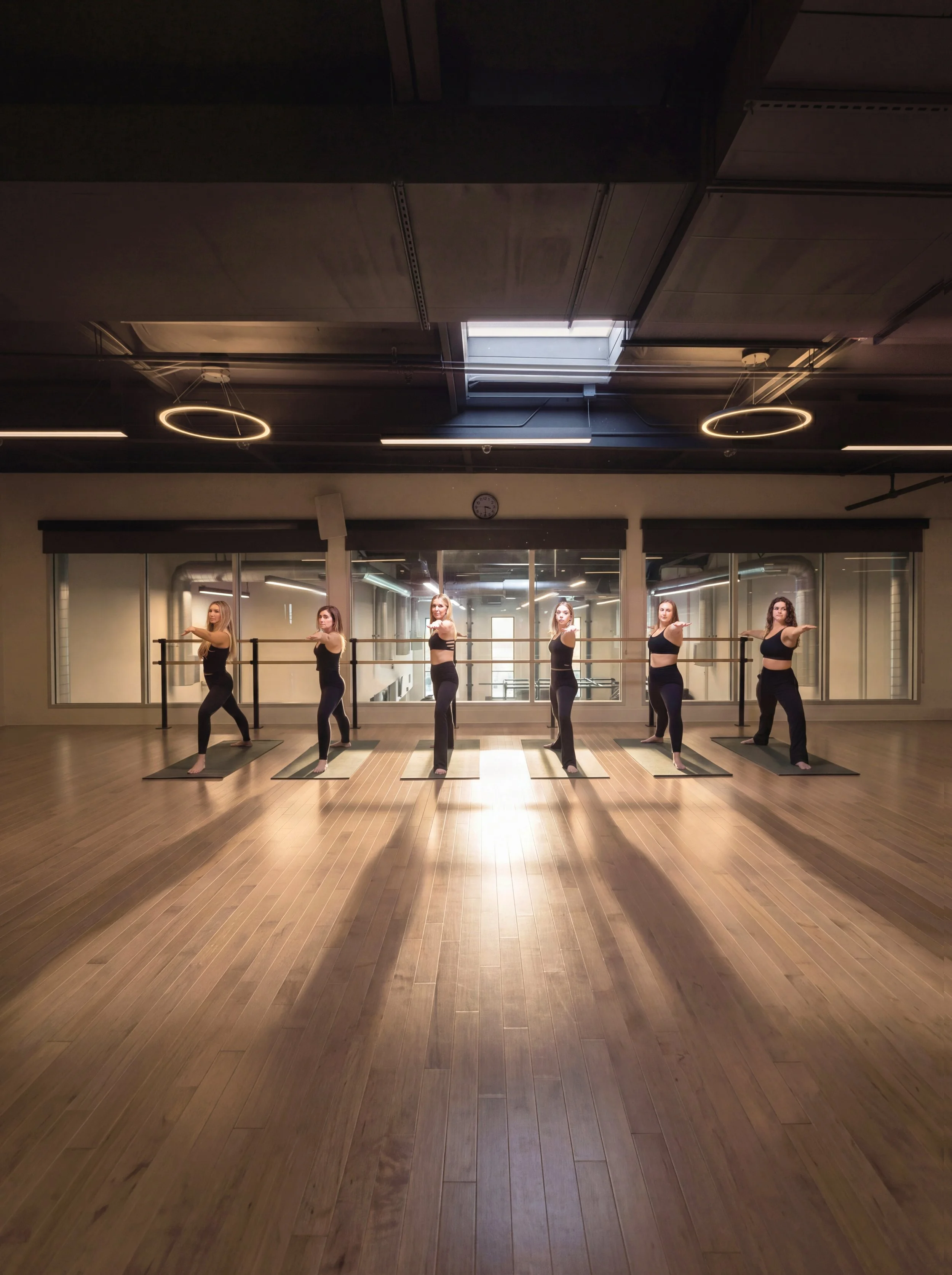 A group of six women practicing ballet in a dance studio with large mirrors, wooden floors, and modern circular light fixtures on the ceiling.