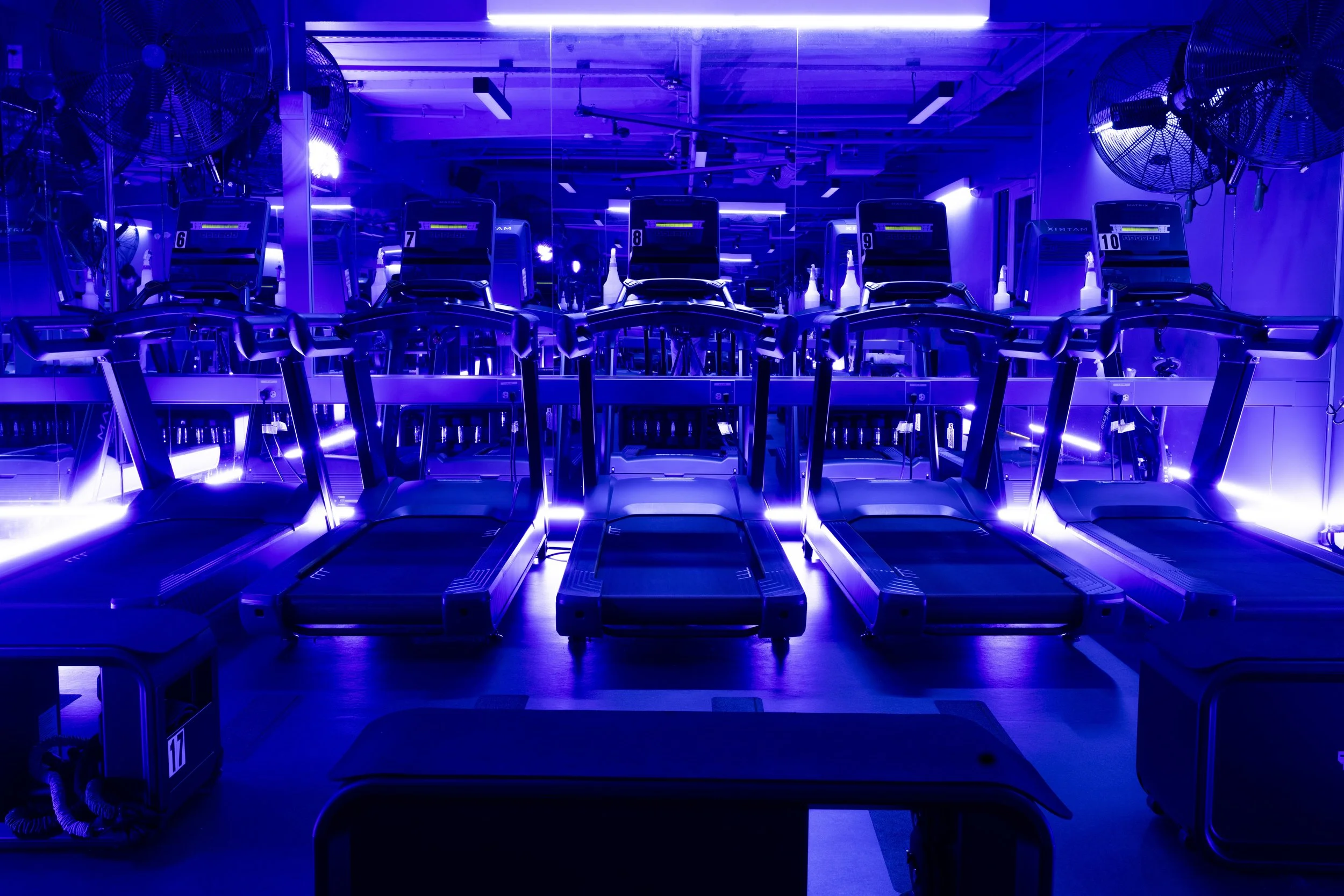 Row of treadmills with blue LED lighting in a gym, mirrored wall behind, large fans mounted on the wall.