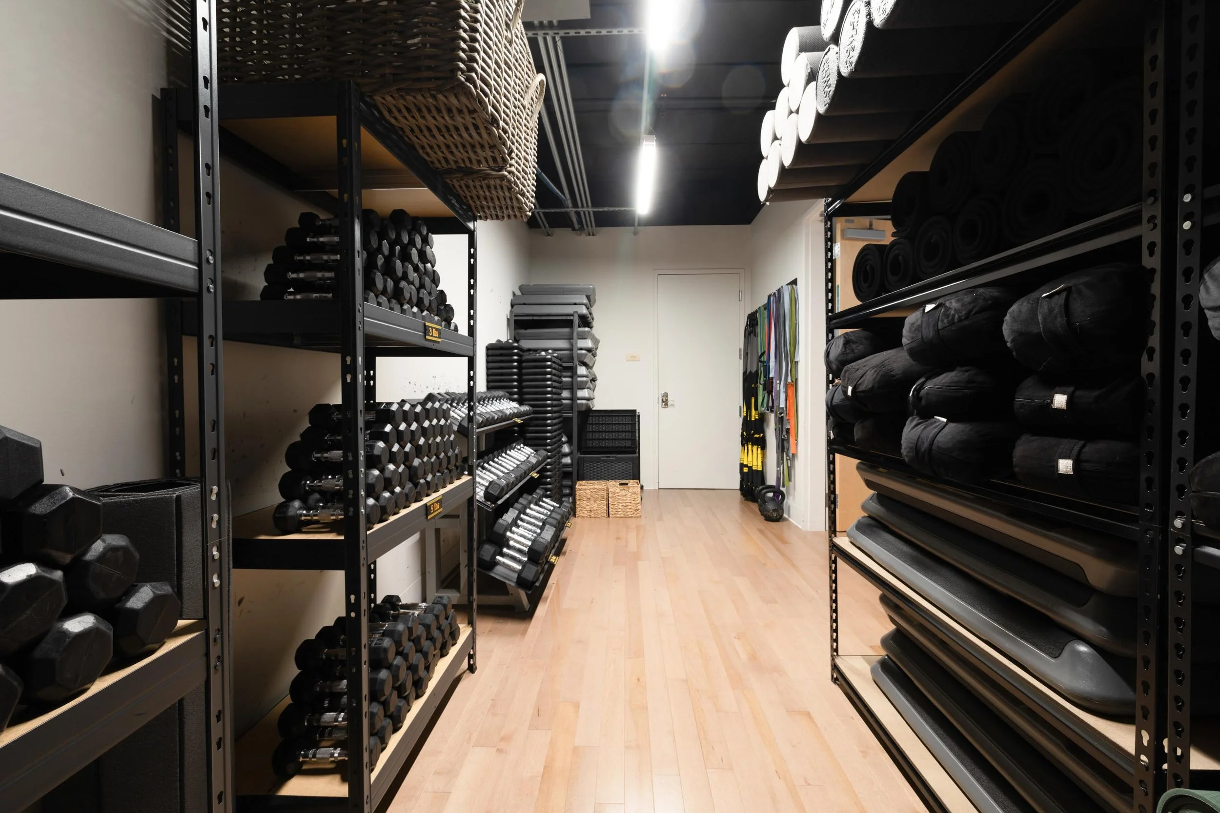 A storage room with black metal shelves holding workout equipment including dumbbells, kettlebells, yoga mats, resistance bands, and foam rollers.