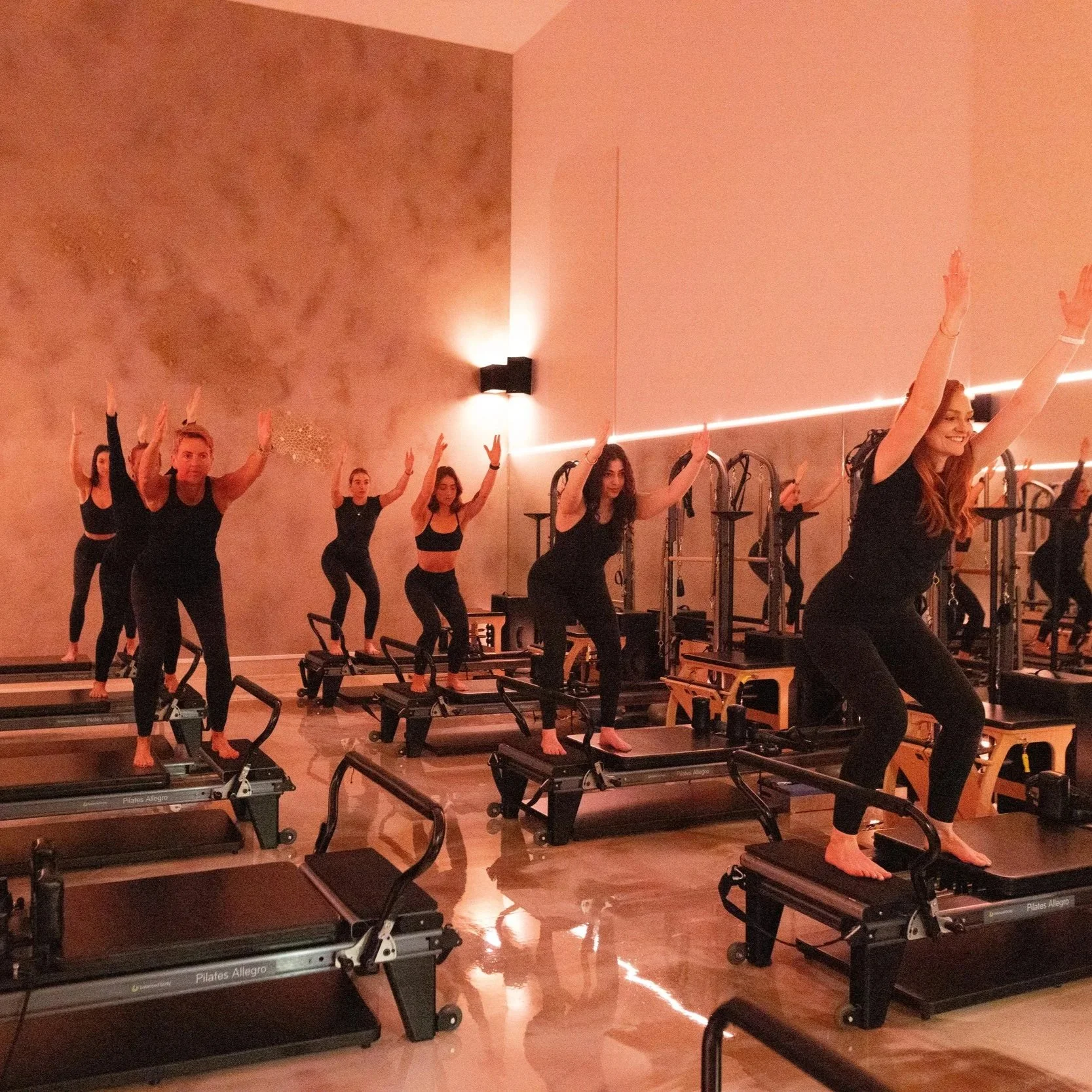 A group of six women and one man participating in a Pilates class on reformer machines in a dimly lit studio with warm lighting, some standing with arms raised and smiling.