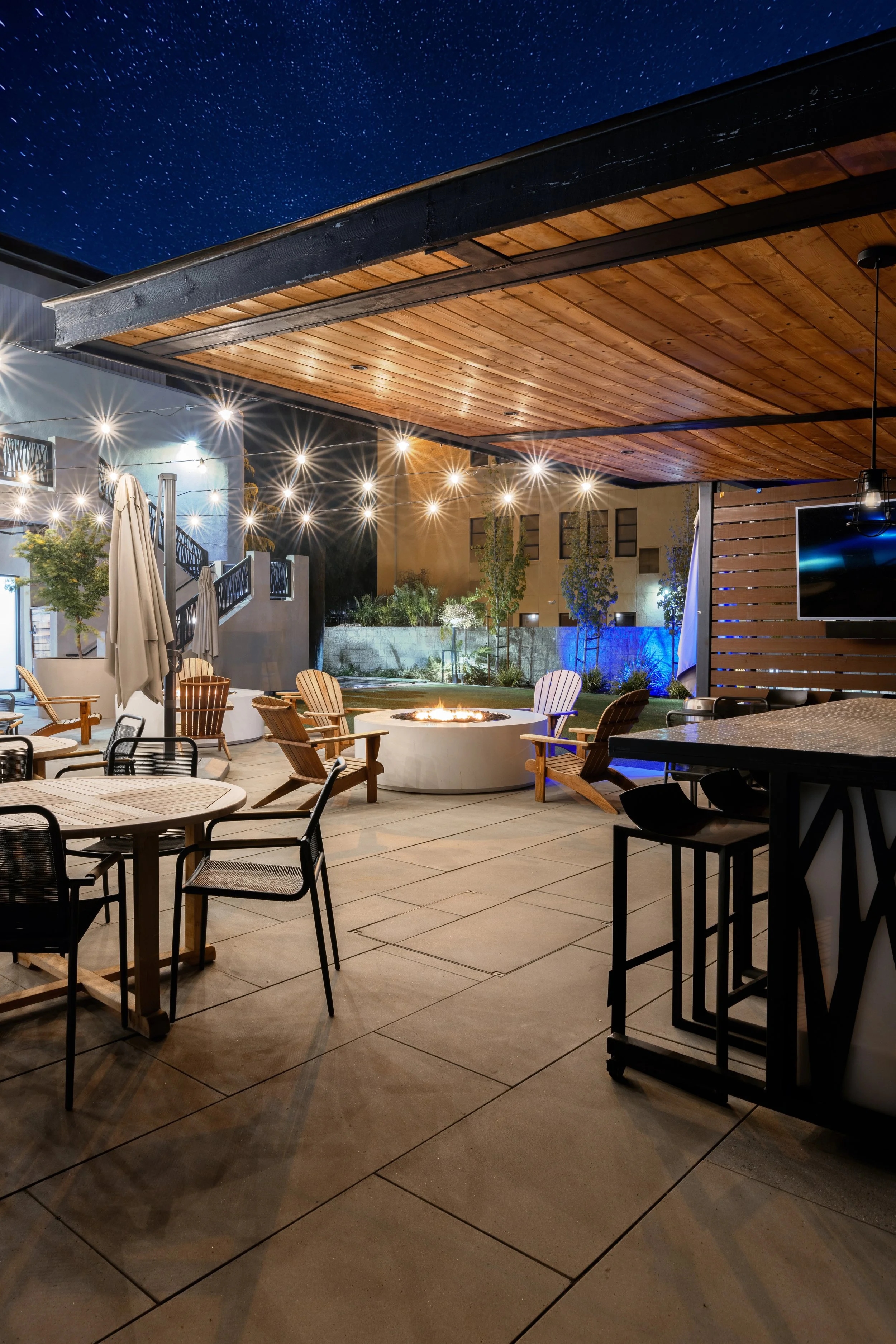 An outdoor patio area at night with string lights, seating, a fire pit, a wooden ceiling, and a starry sky.