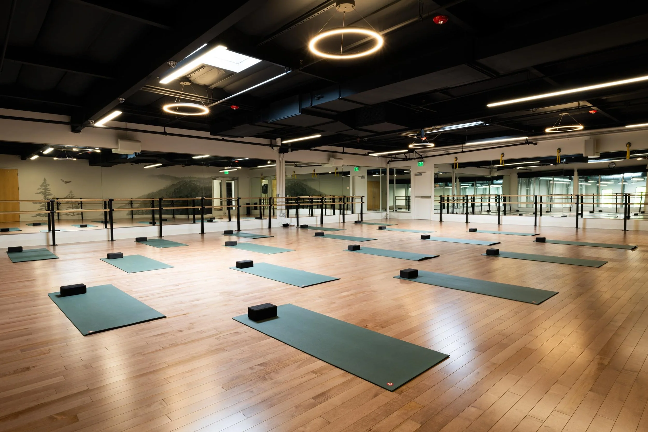 A spacious yoga studio with wooden floors, yoga mats, and blocks arranged in rows. The room has mirrored walls, decorative wall murals of nature scenes, and modern lighting fixtures hanging from the black ceiling.