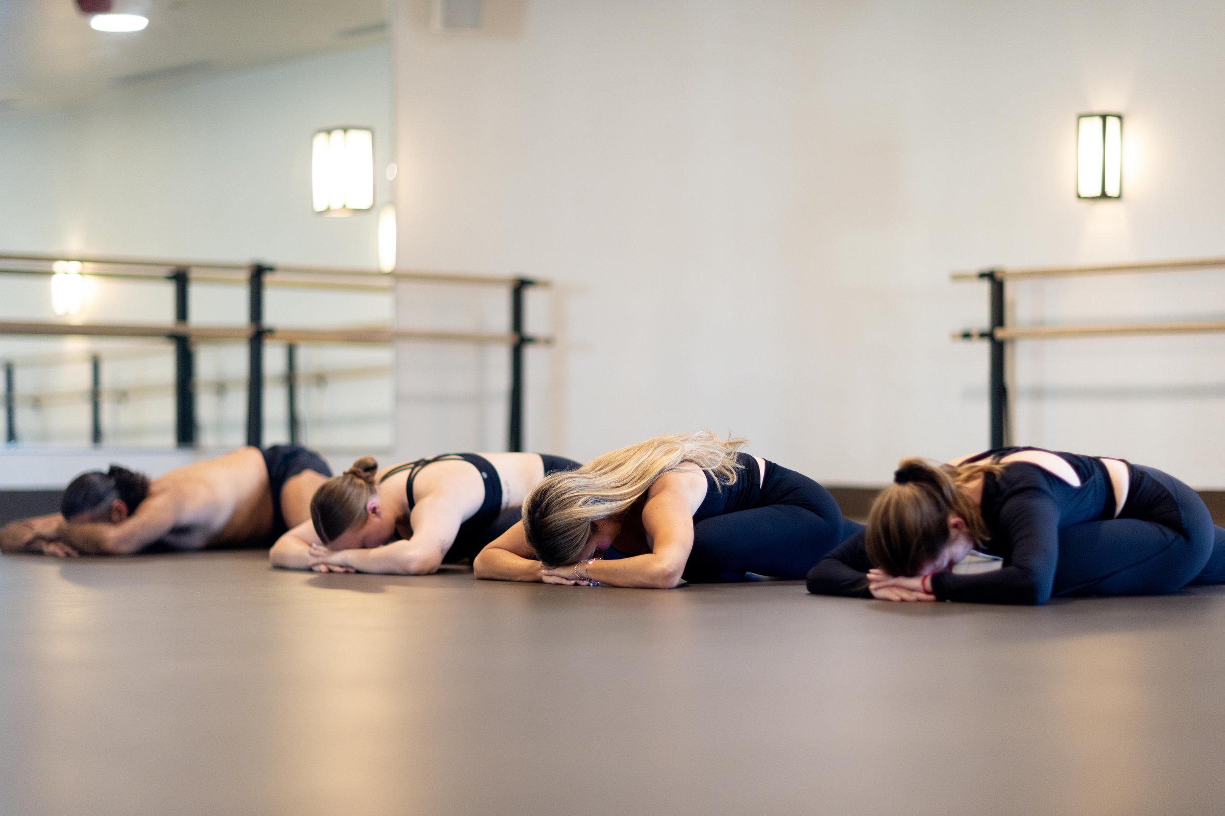 Group of people practicing yoga or meditation in a studio, lying face down in a pose, with ballet barres and wall lights in the background.