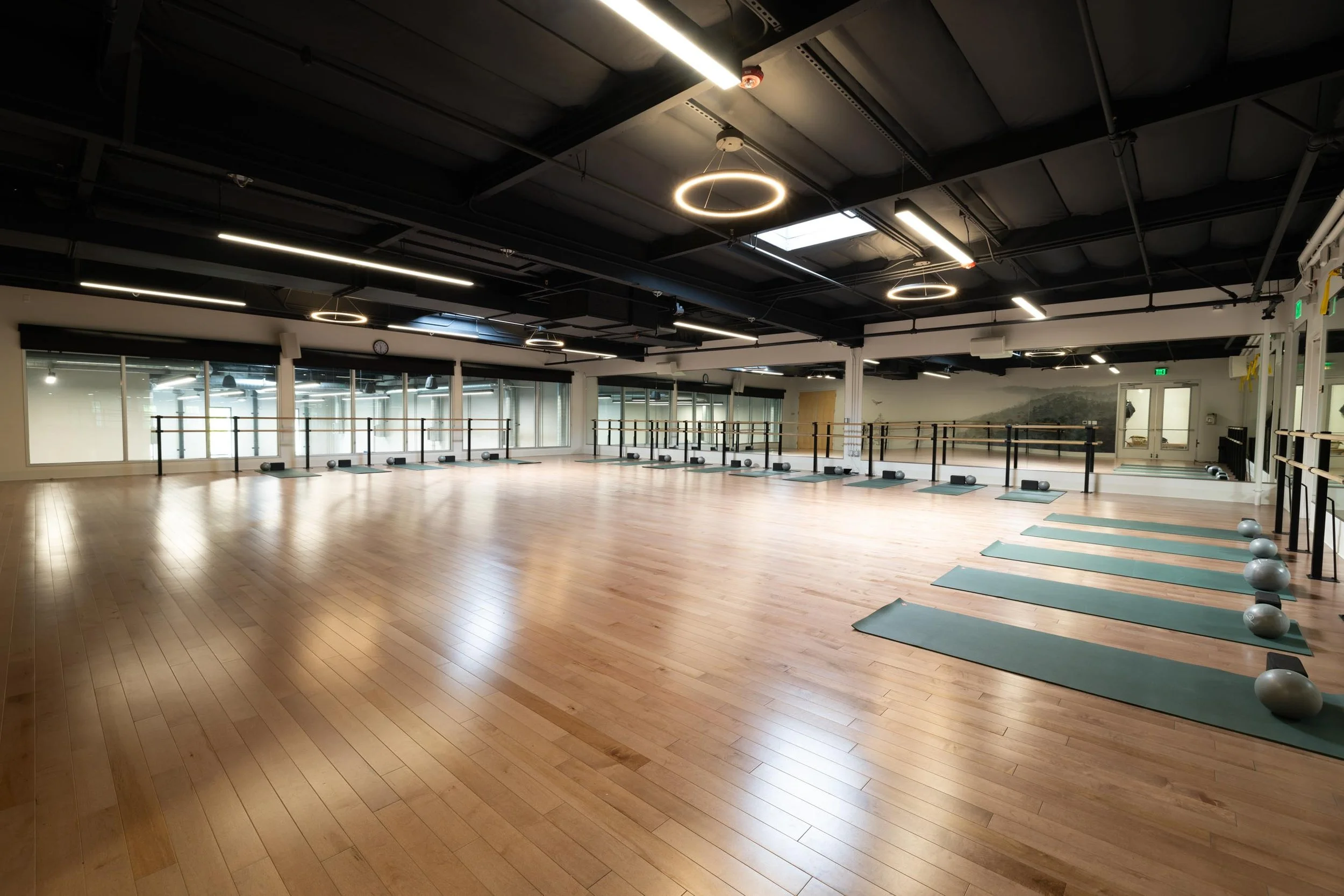 Empty spacious workout studio with wooden floors, large mirrors, and windows. Exercise mats and large fitness balls are arranged near the mirrors. Modern ceiling lights and circular pendant lights are installed, with some natural light coming from skylights.
