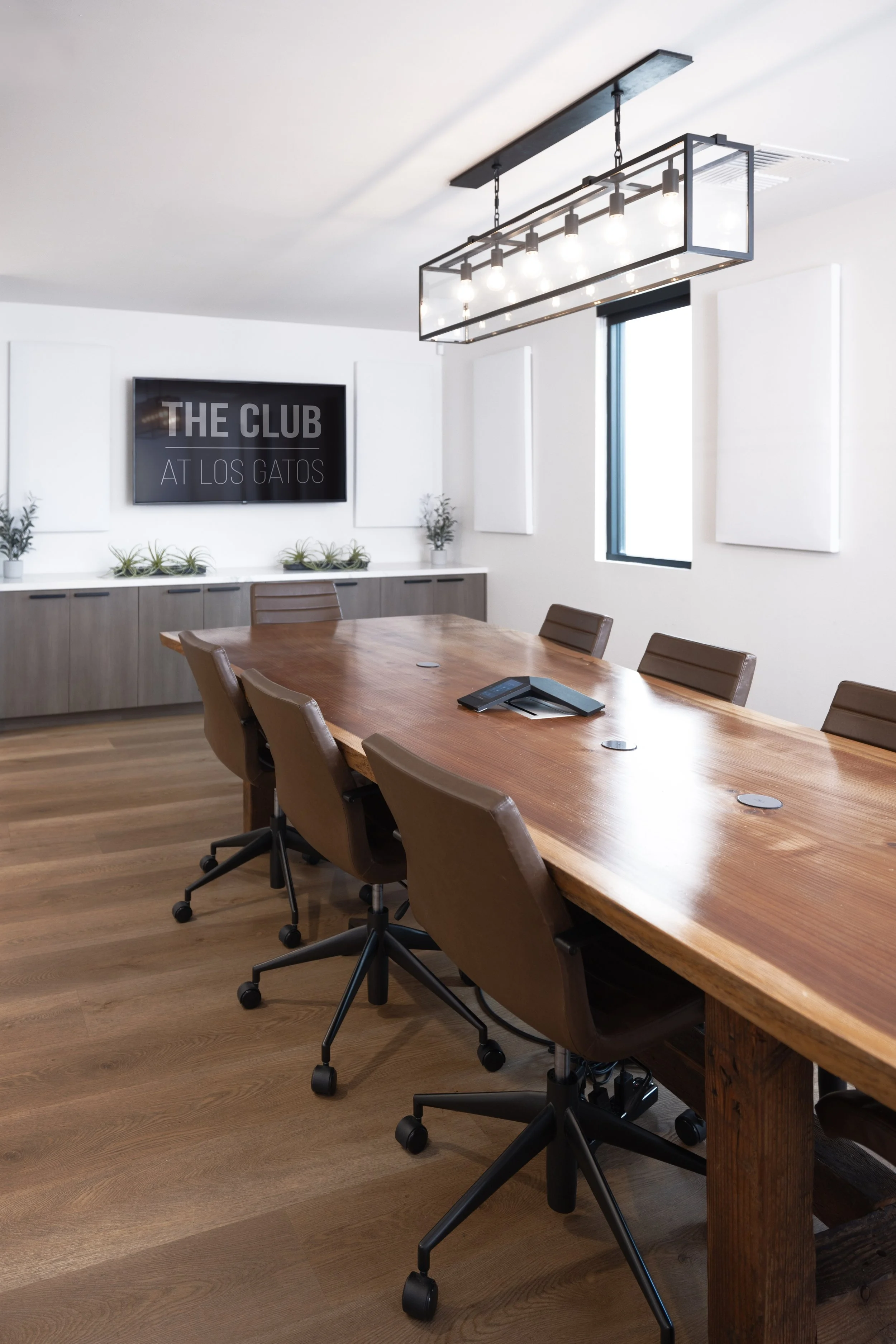 A modern conference room with a long wooden table, seven office chairs, and a rectangular light fixture hanging from the ceiling. The room features white walls, a small window, and some green plants on a cabinet.