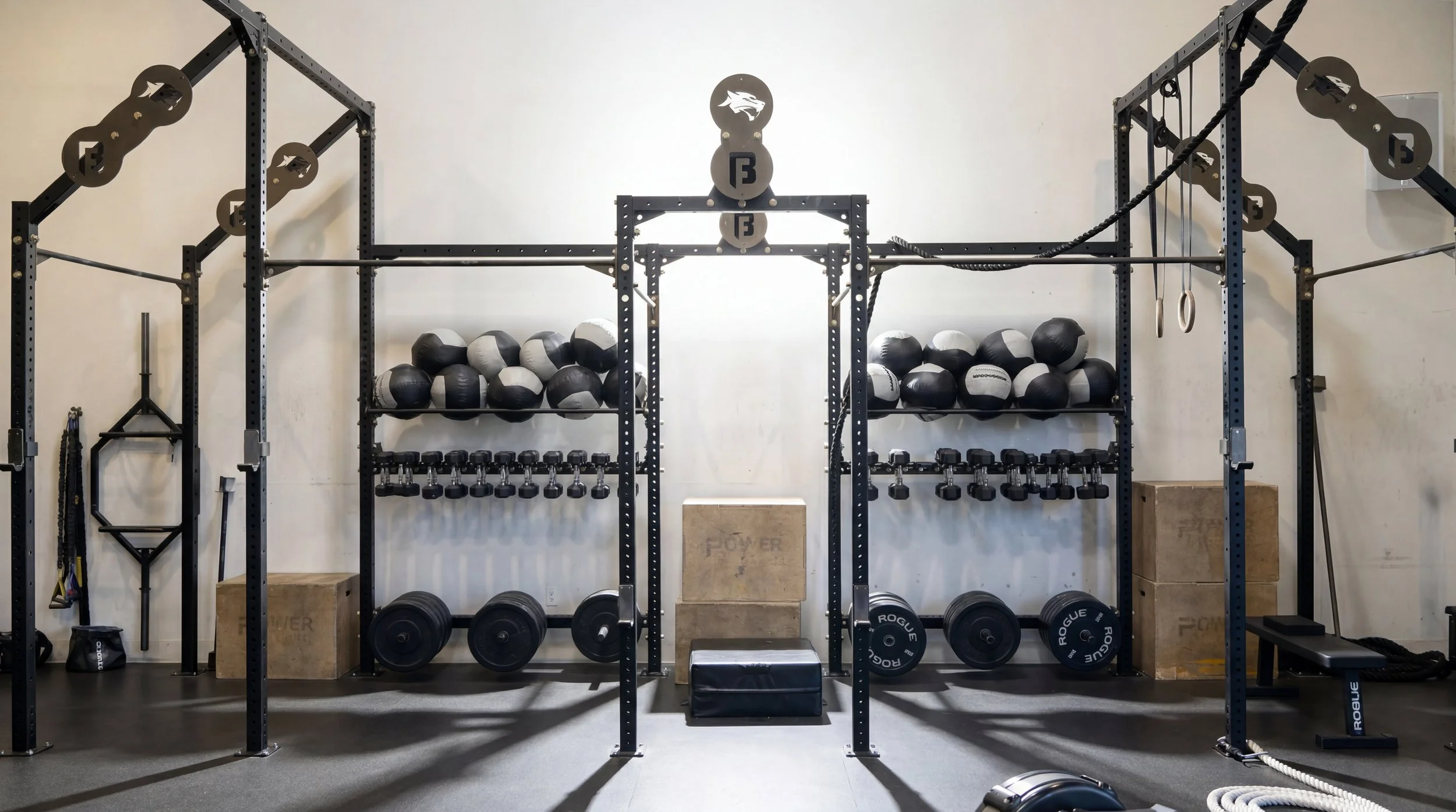 A fitness gym setup with a large black metal power rack, black and white medicine balls on shelves, dumbbells, weight plates, and various workout equipment organized on the wall and floor.