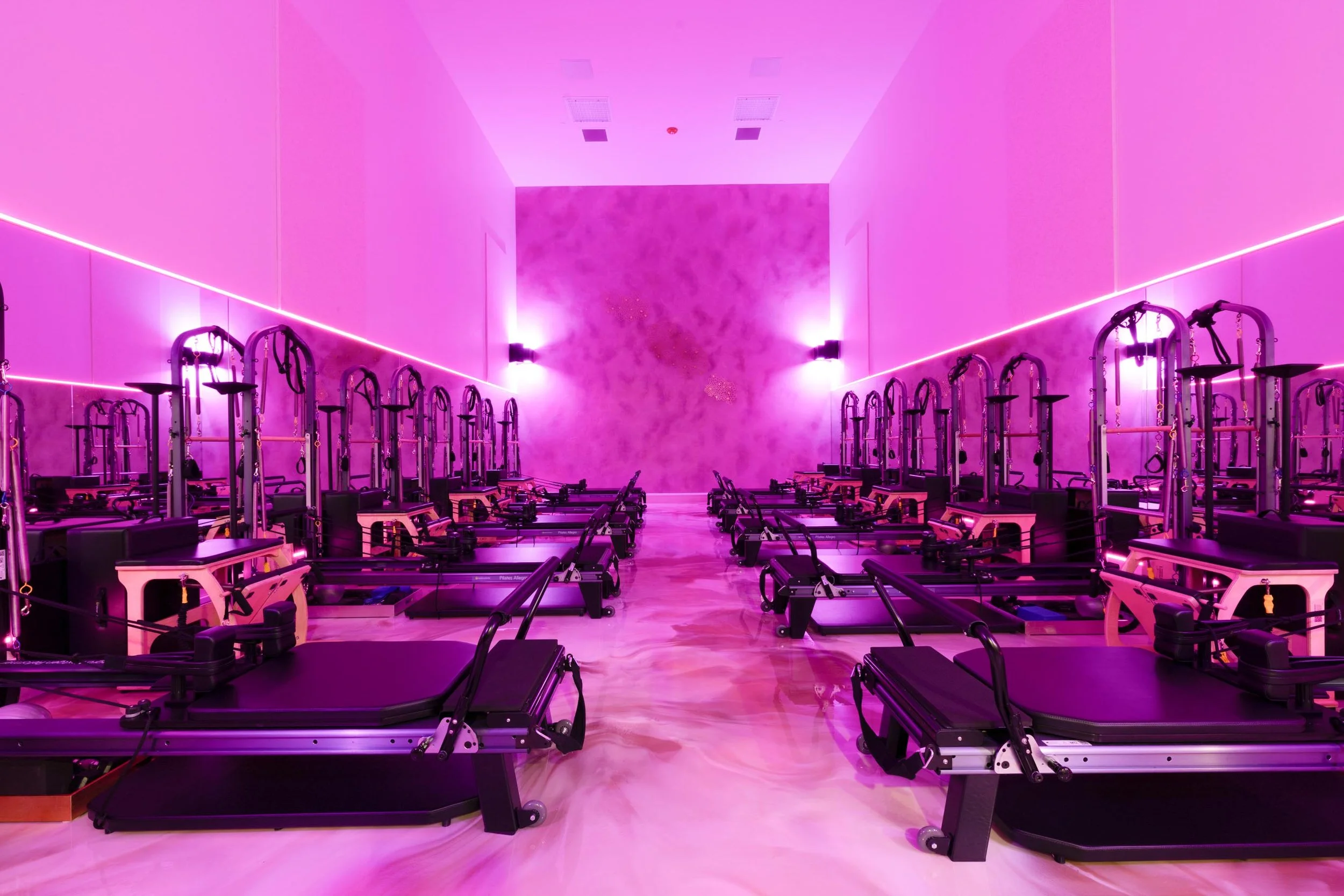 A room with pink lighting and Pilates reformer machines lined up along the walls.