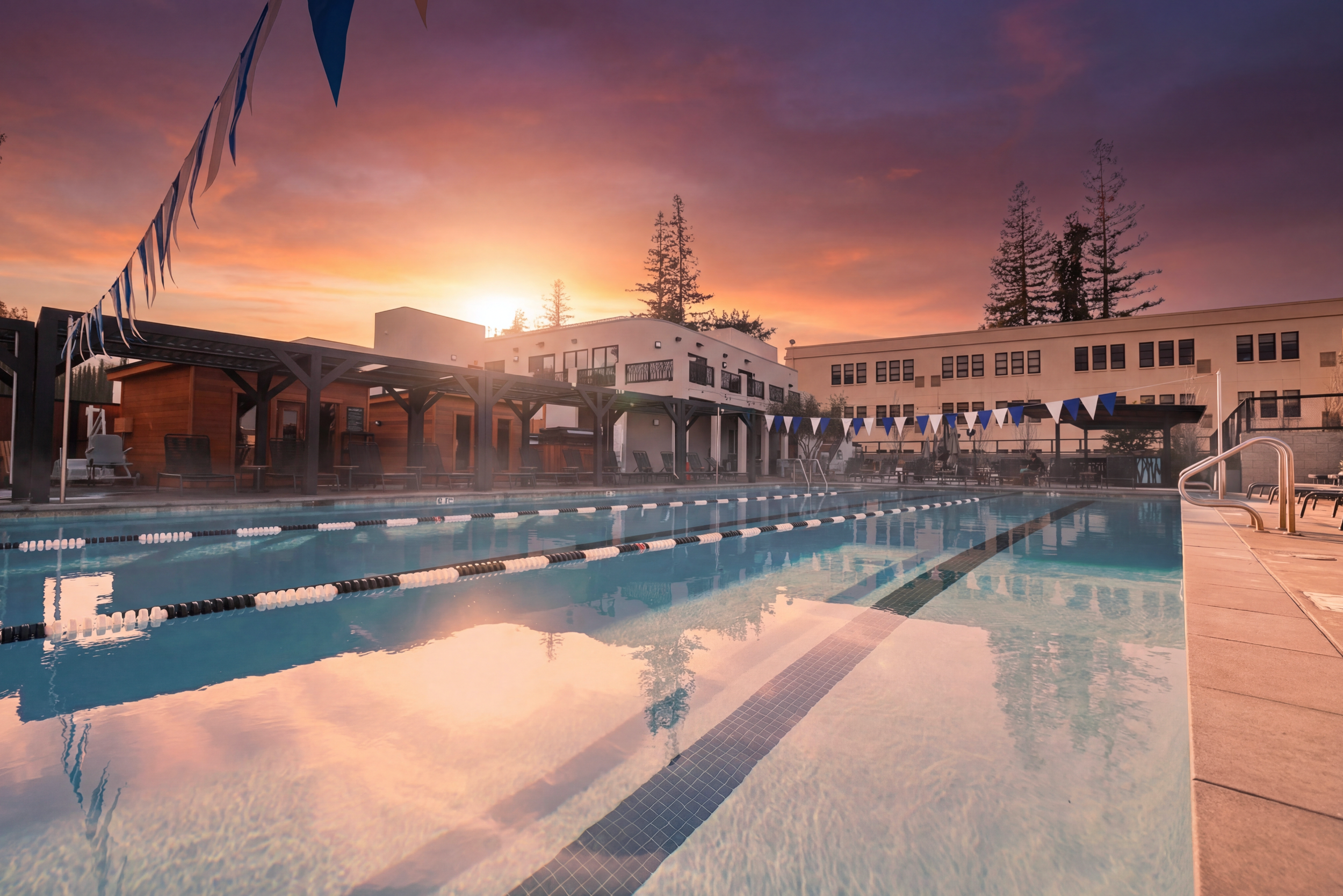 An outdoor swimming pool at sunset with lounge chairs, a wooden cabana, white and blue triangular flags, and buildings in the background, surrounded by tall trees.