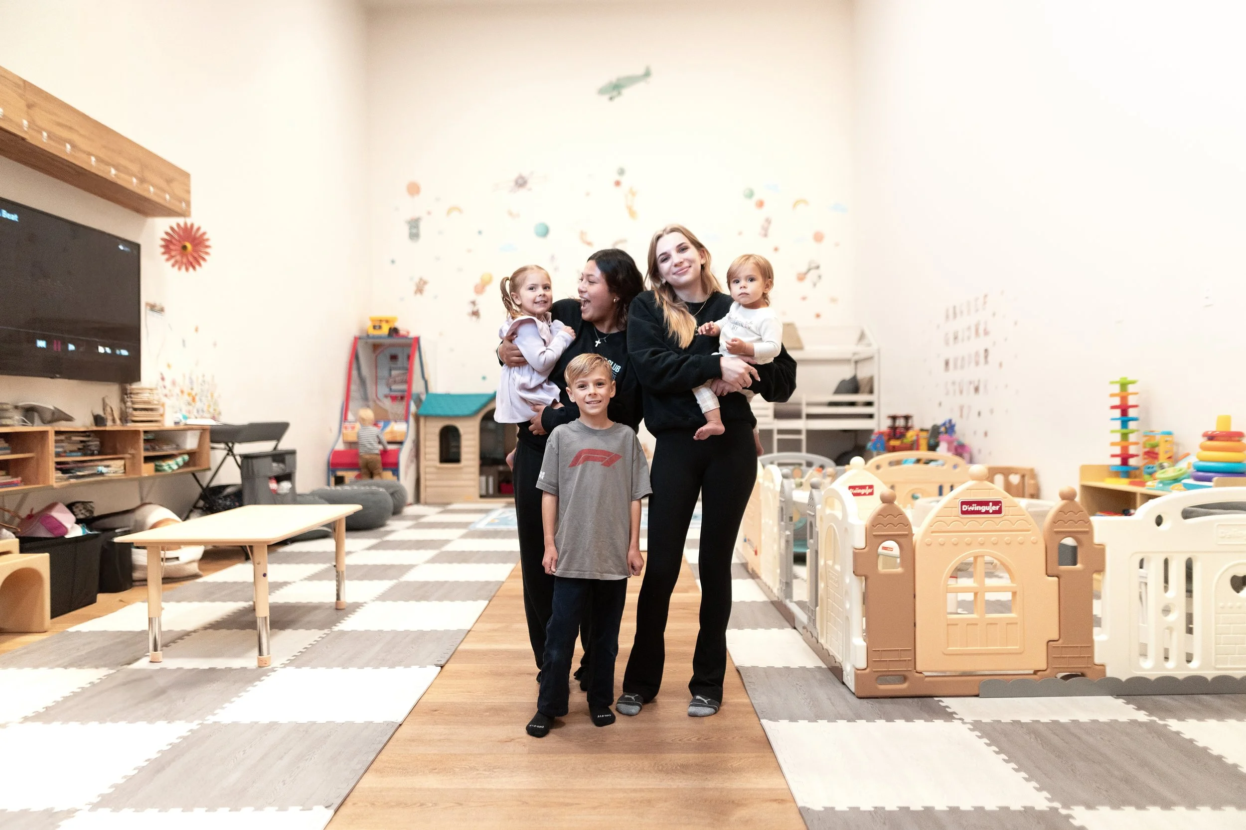 Childcare Attendants and 3 young children, various ages standing in a playroom filled with toys and play structures. All are happy, safe, content and smiling.