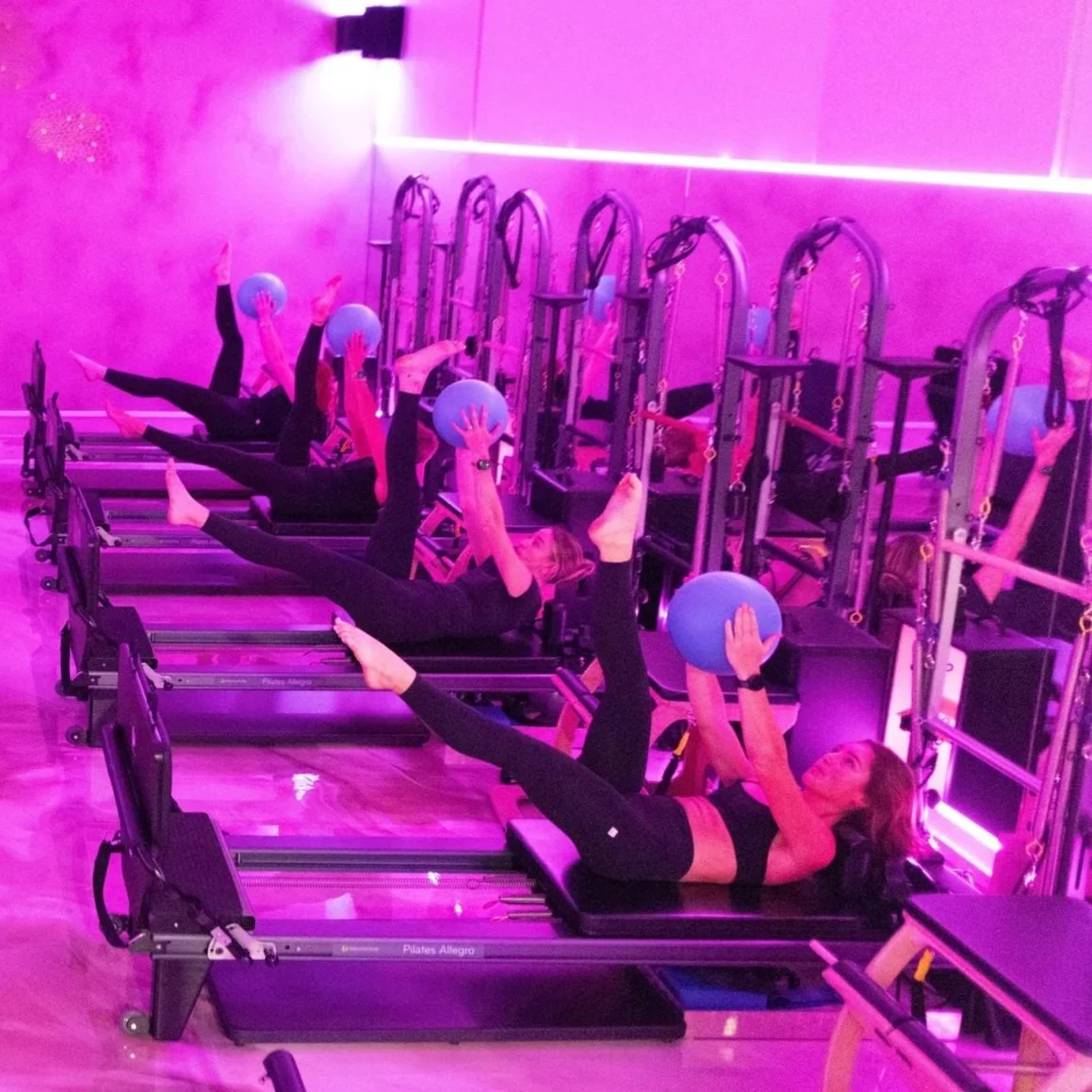 Group of women exercising with small blue balls on Pilates reformer machines in a  pink-lit fitness studio.