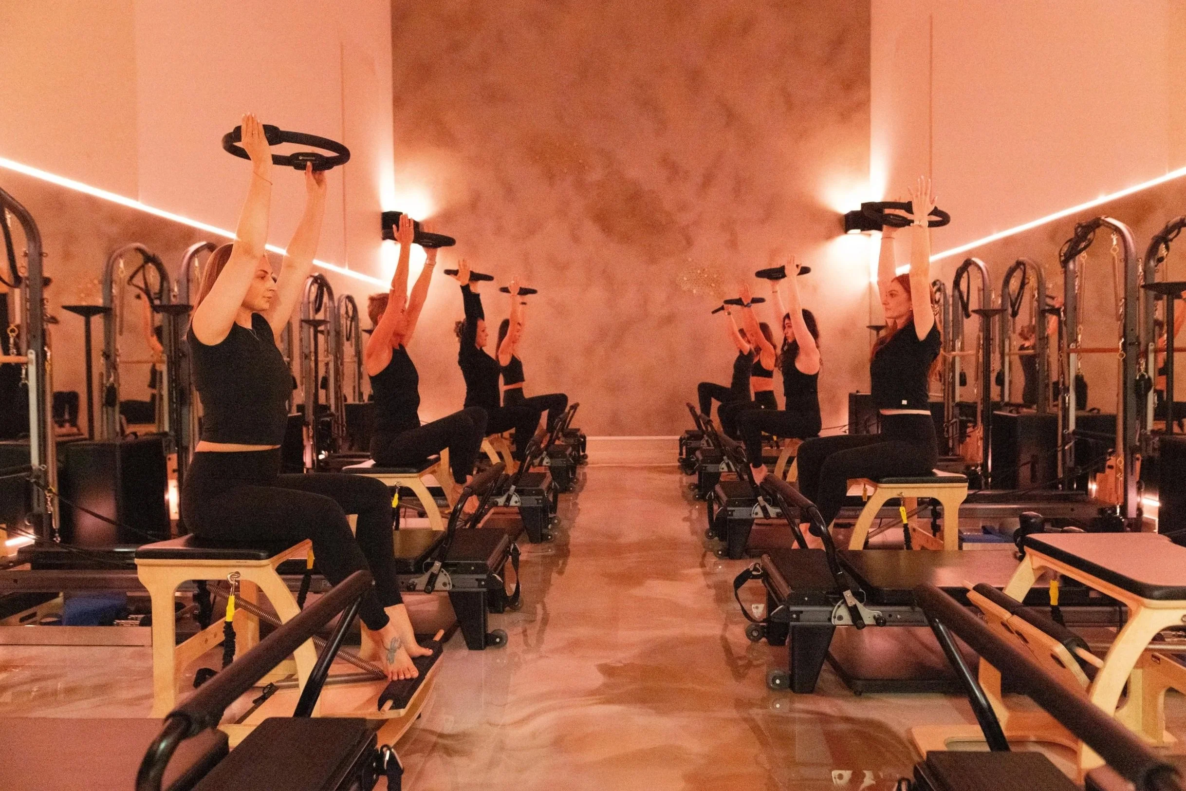 A group of women participating in a Pilates class, using reformer machines in a dimly lit studio with modern lighting and a textured beige wall.