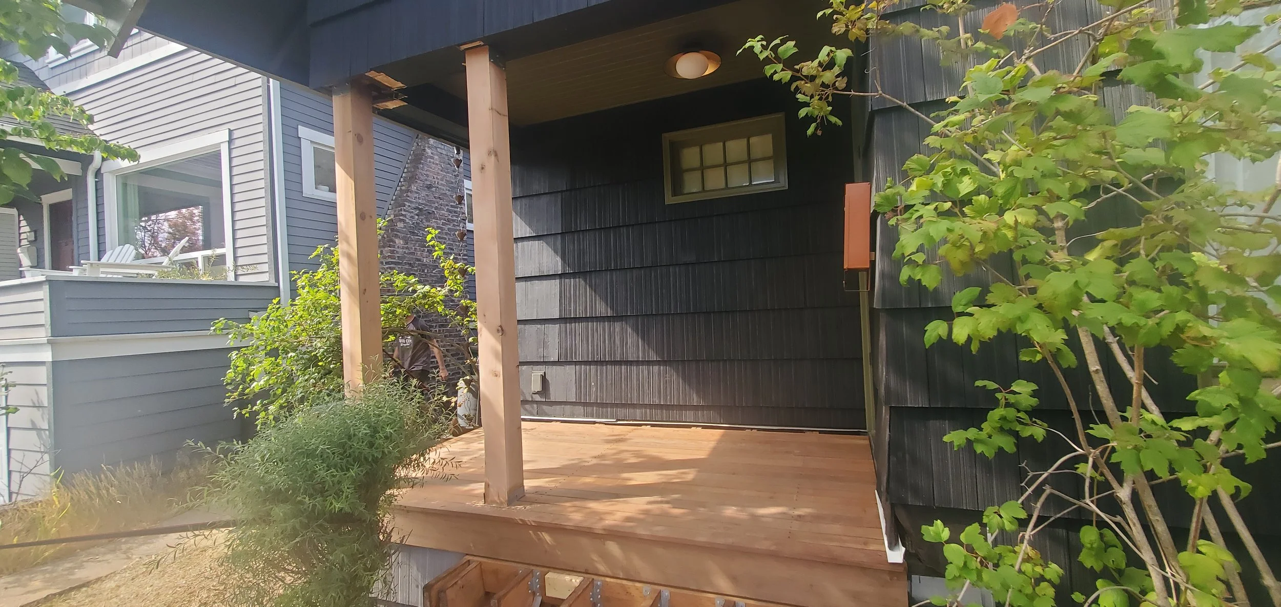 Newly constructed porch with wooden deck and support beams attached to the side of a house with black exterior siding, surrounded by green foliage.