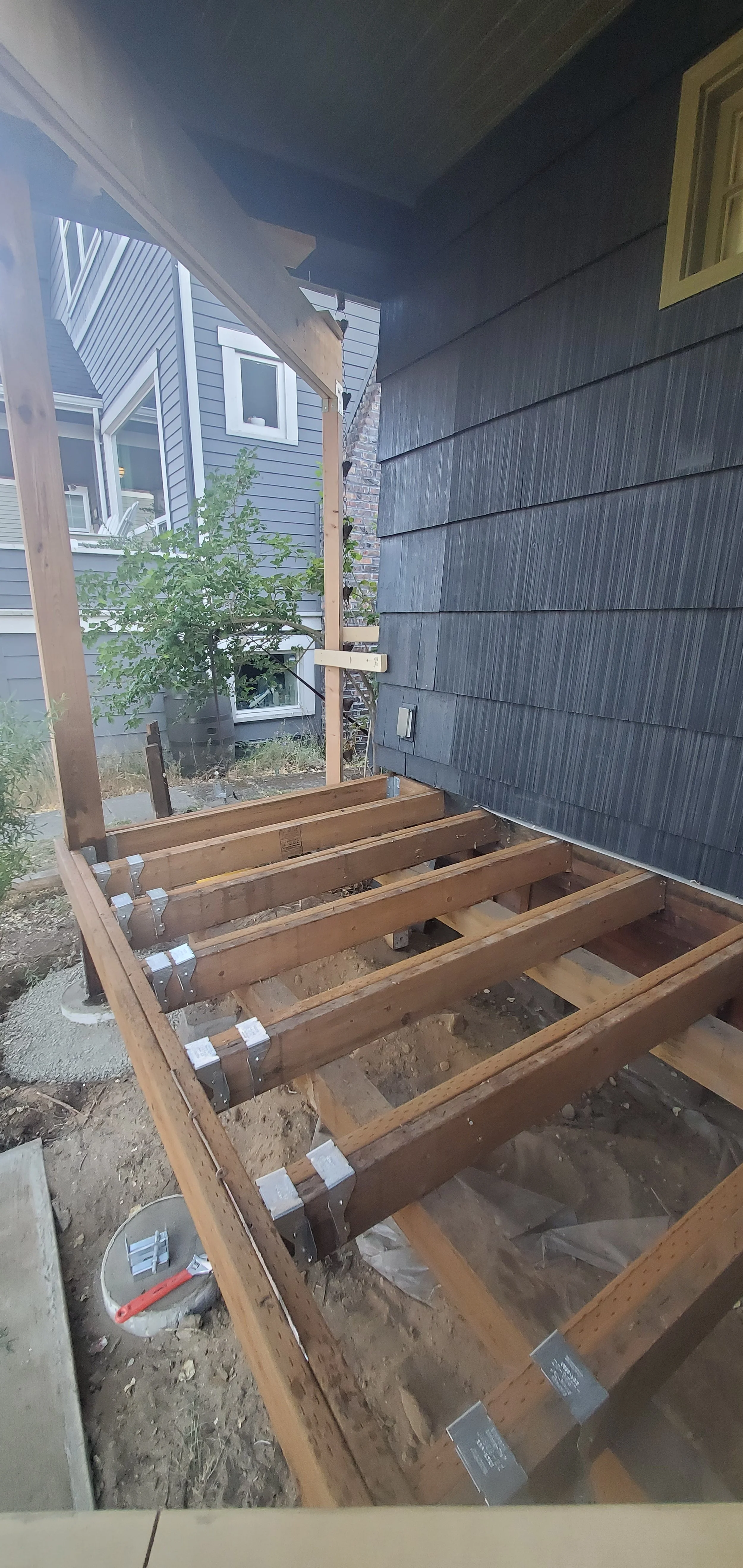 Construction of a deck or porch with wooden beams and supports in a backyard, next to a house with black siding.