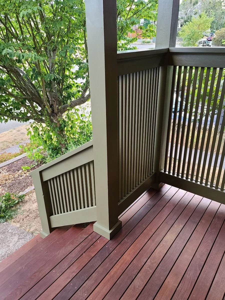 Close-up of a wooden porch with brown flooring, a beige and dark brown railing, and a view of a tree and greenery outside.