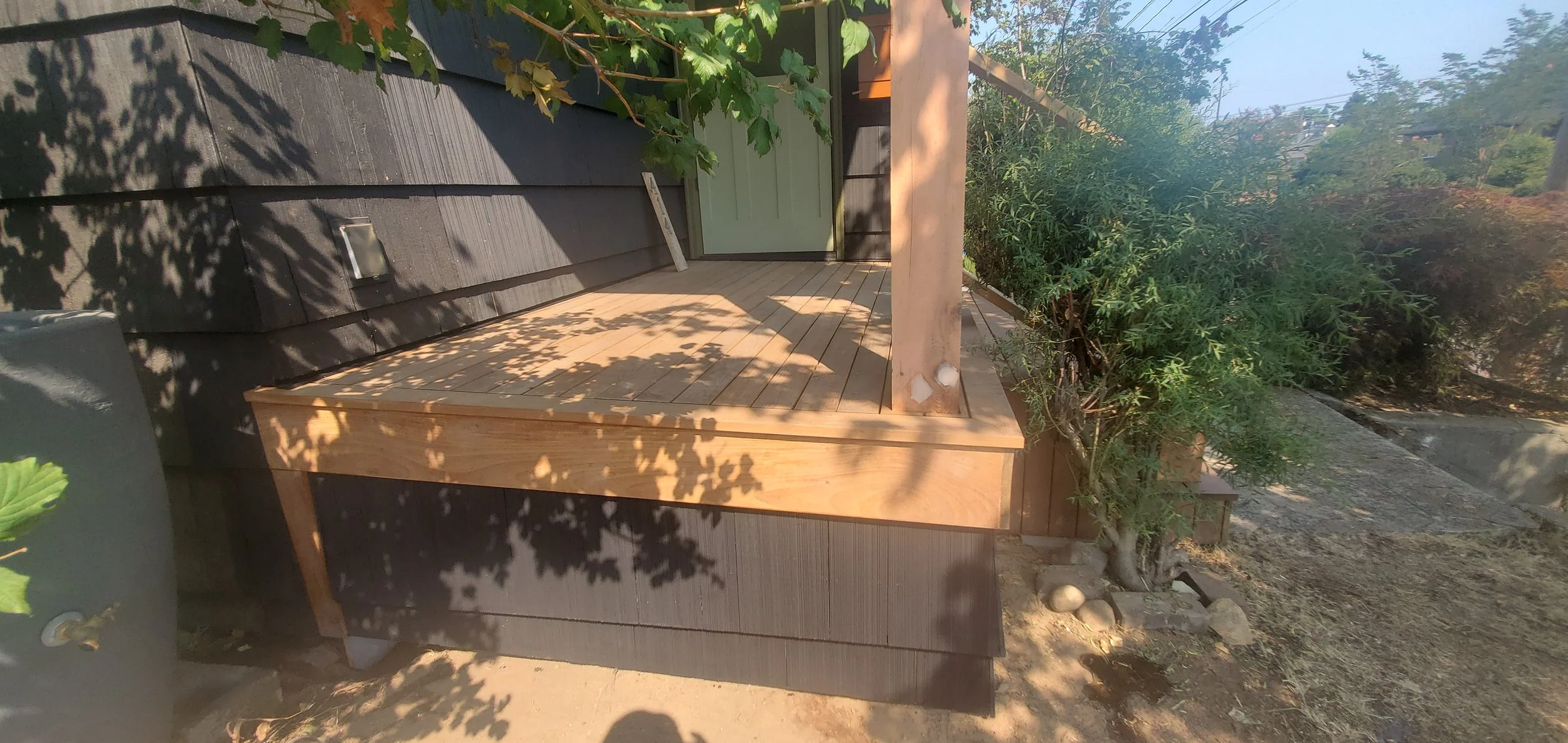 A small wooden deck attached to the side of a dark gray house, with a white door. The deck is surrounded by green plants and trees, casting shadows on the surface. There are rocks and soil on the ground nearby.
