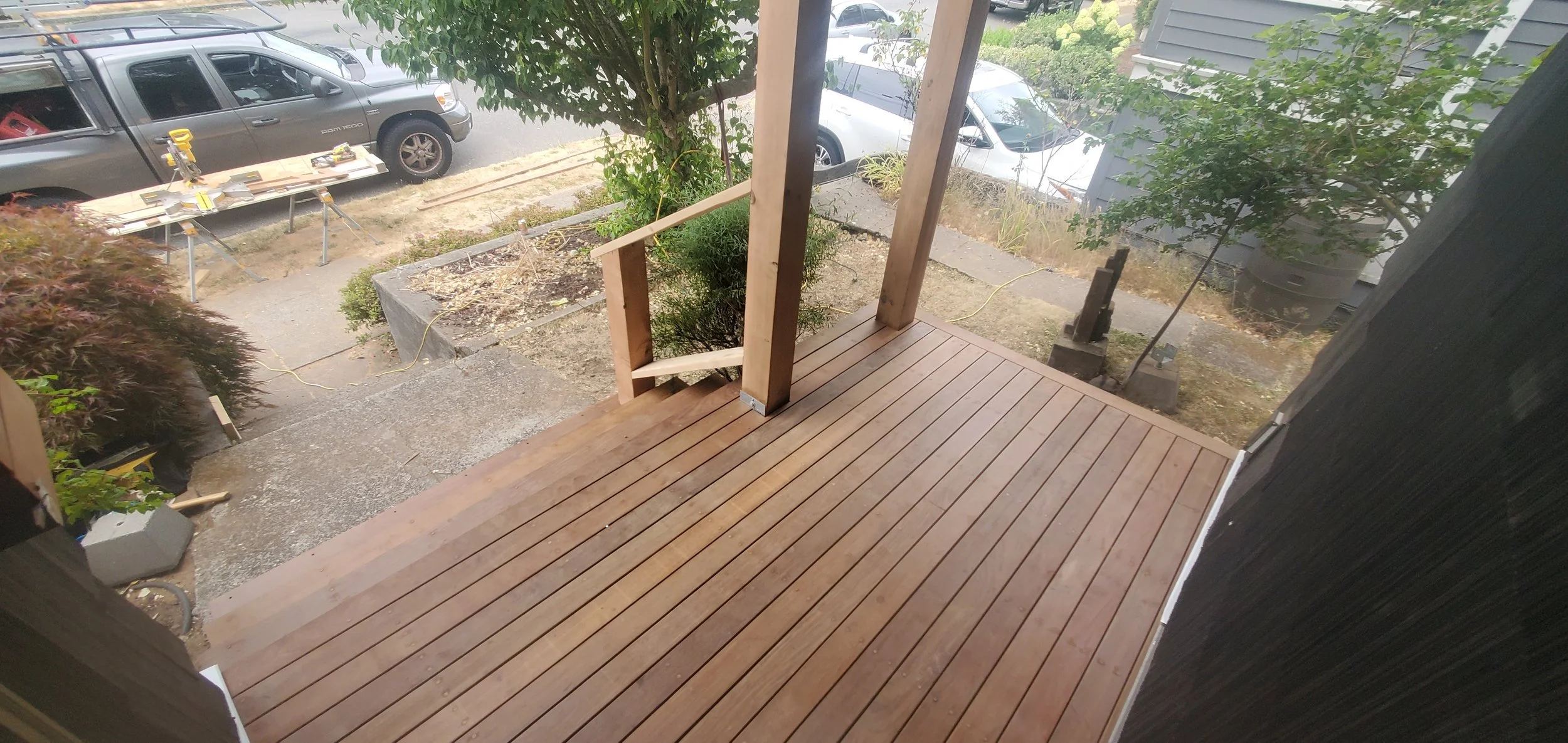 View from a porch showing wooden flooring, unfinished railing, nearby plants, and a construction area with parked cars in the background.