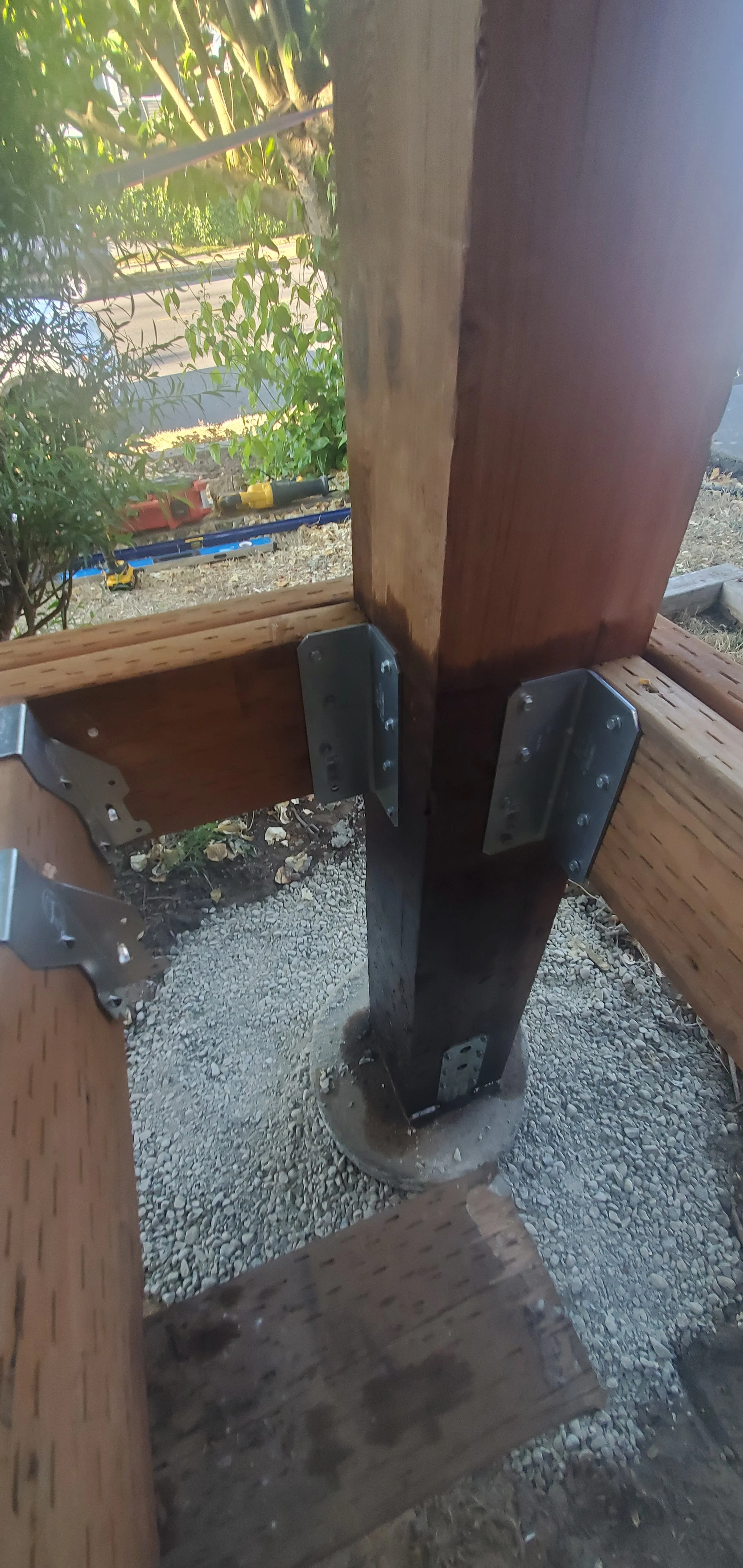 Close-up view of a wooden outdoor structure under construction with metal brackets connecting wooden beams to a vertical post, on a gravel ground.