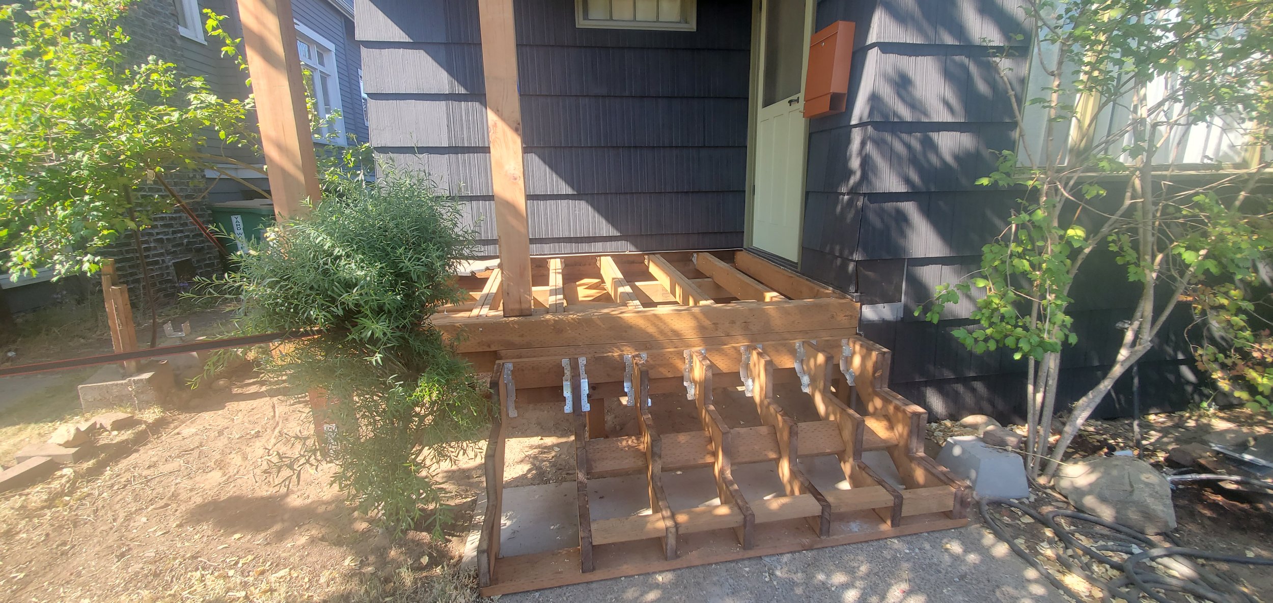 Front porch under construction with wooden framing, installed on a house with blue shingle siding. A white door is visible with a small orange mailbox beside it. There is a bush to the left, and a tree with green leaves on the right. Construction mat