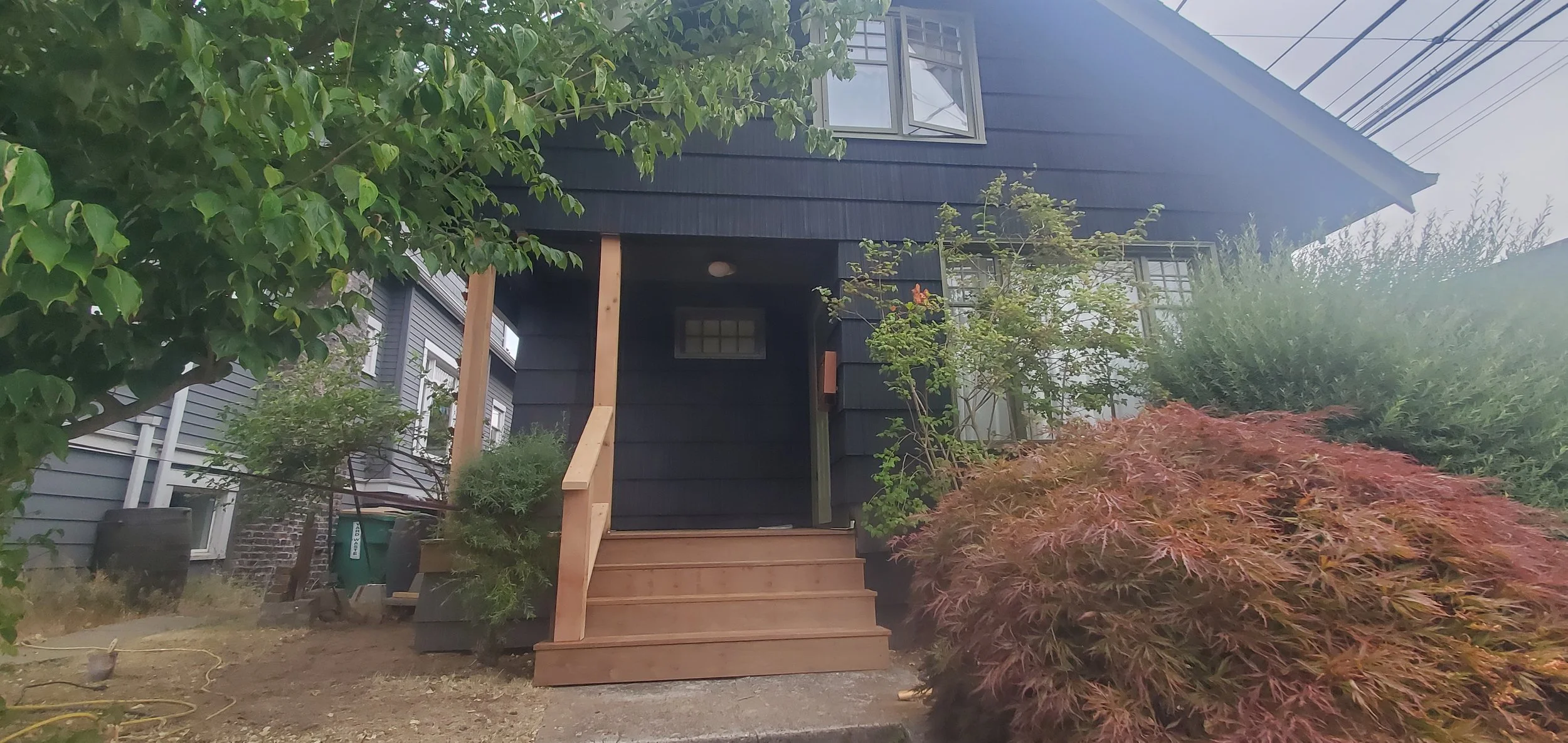 A house with a black exterior and a small front porch under construction, surrounded by trees, bushes, and gardening tools.