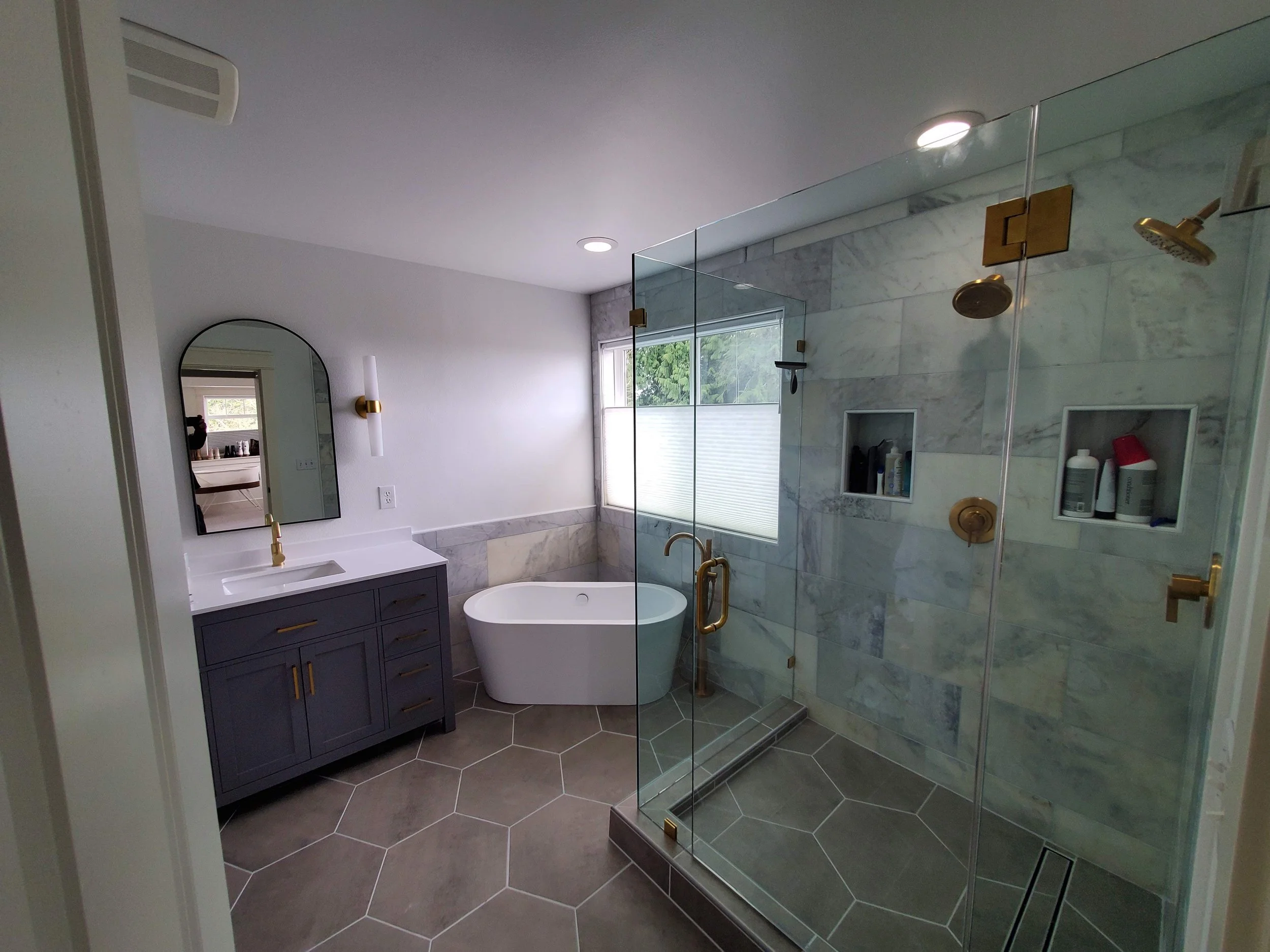 Modern bathroom with a glass shower enclosure, a freestanding bathtub, a dark vanity with a white countertop and gold fixtures, hexagonal tile flooring, a large window, a mirror, and wall-mounted light fixtures.