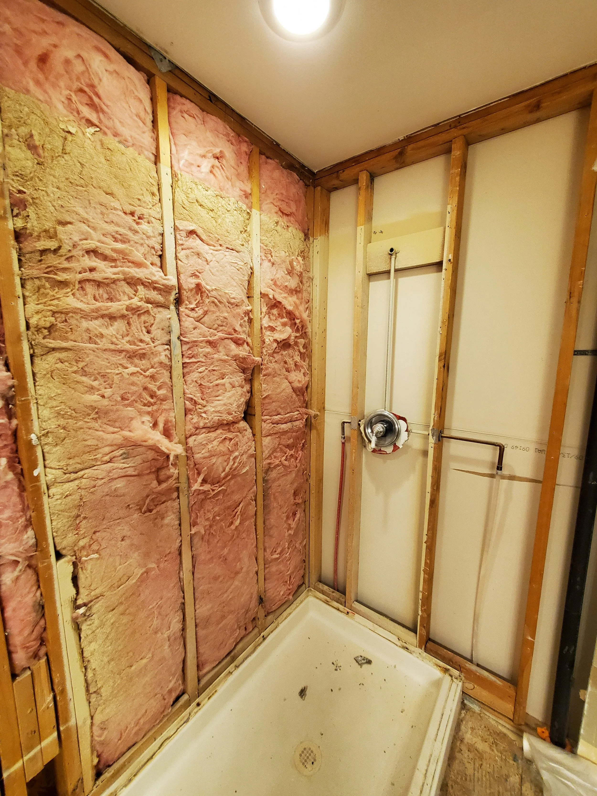 Bathroom renovation in progress, showing exposed pink insulation and wooden framing around a shower base.