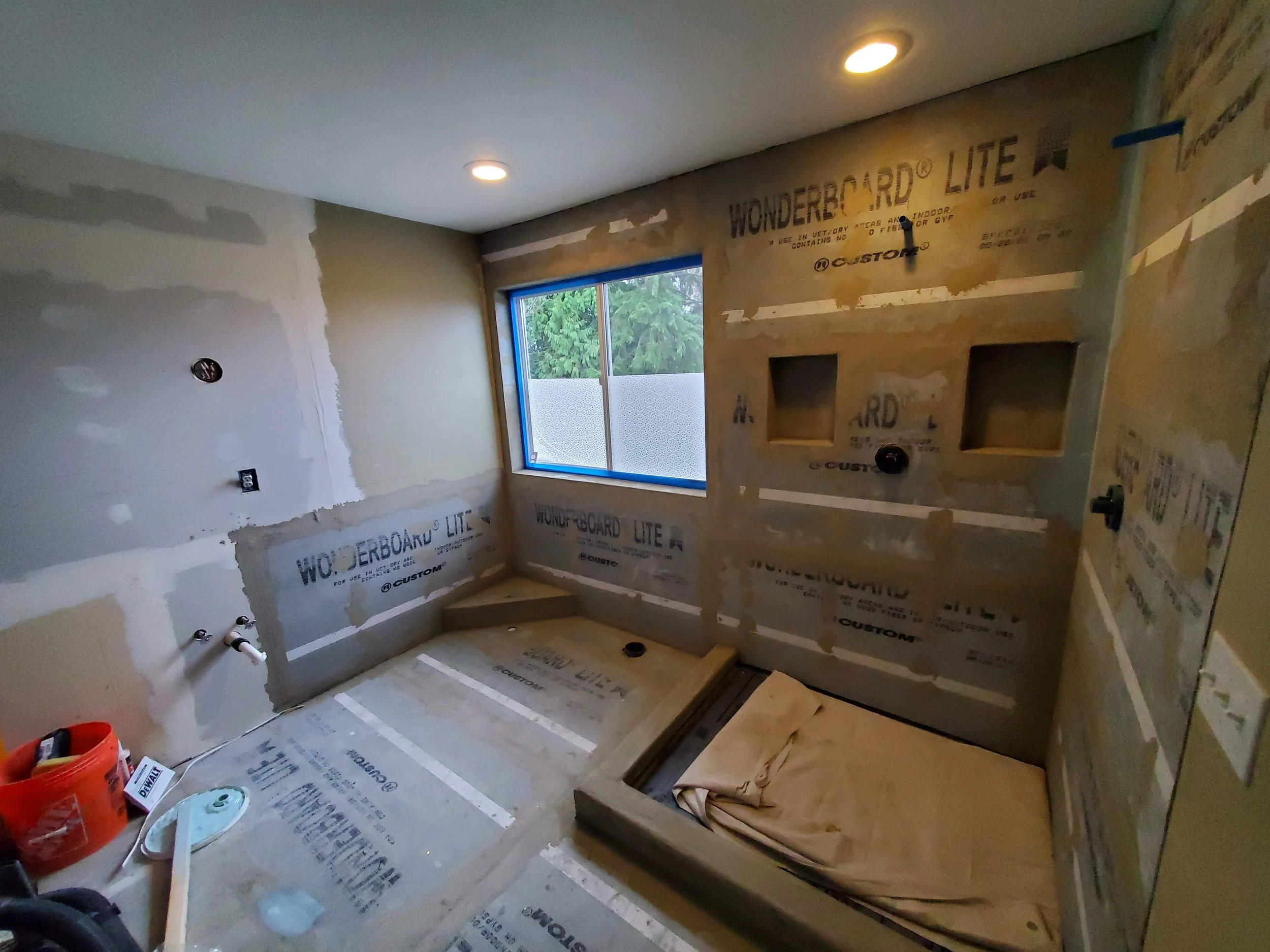 A bathroom under renovation with bare walls and drywall, a window with view of greenery outside, and construction materials on the floor.