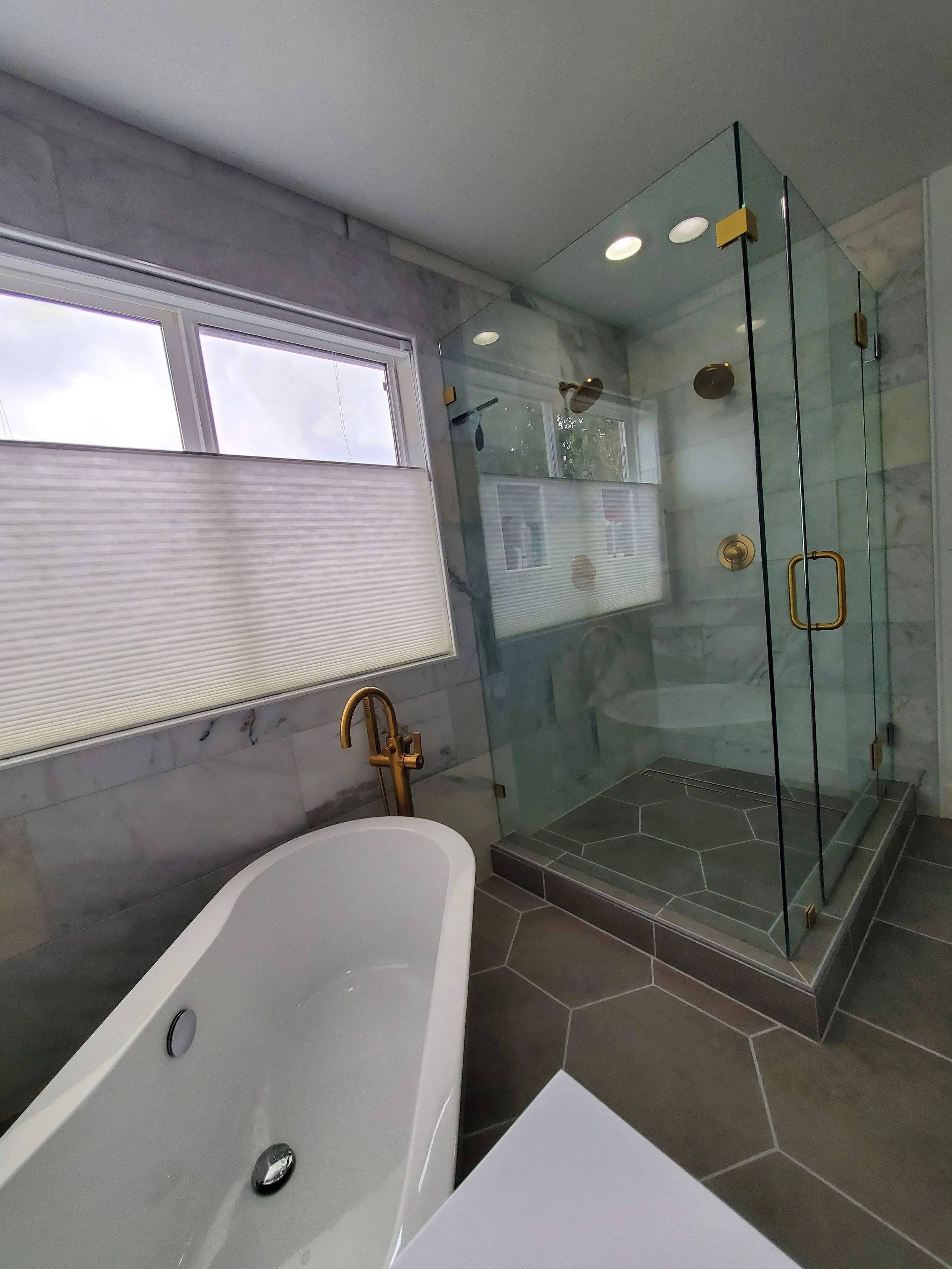 Bathroom with large window, bathtub, and glass-enclosed shower, featuring brass fixtures and gray tile flooring and walls.