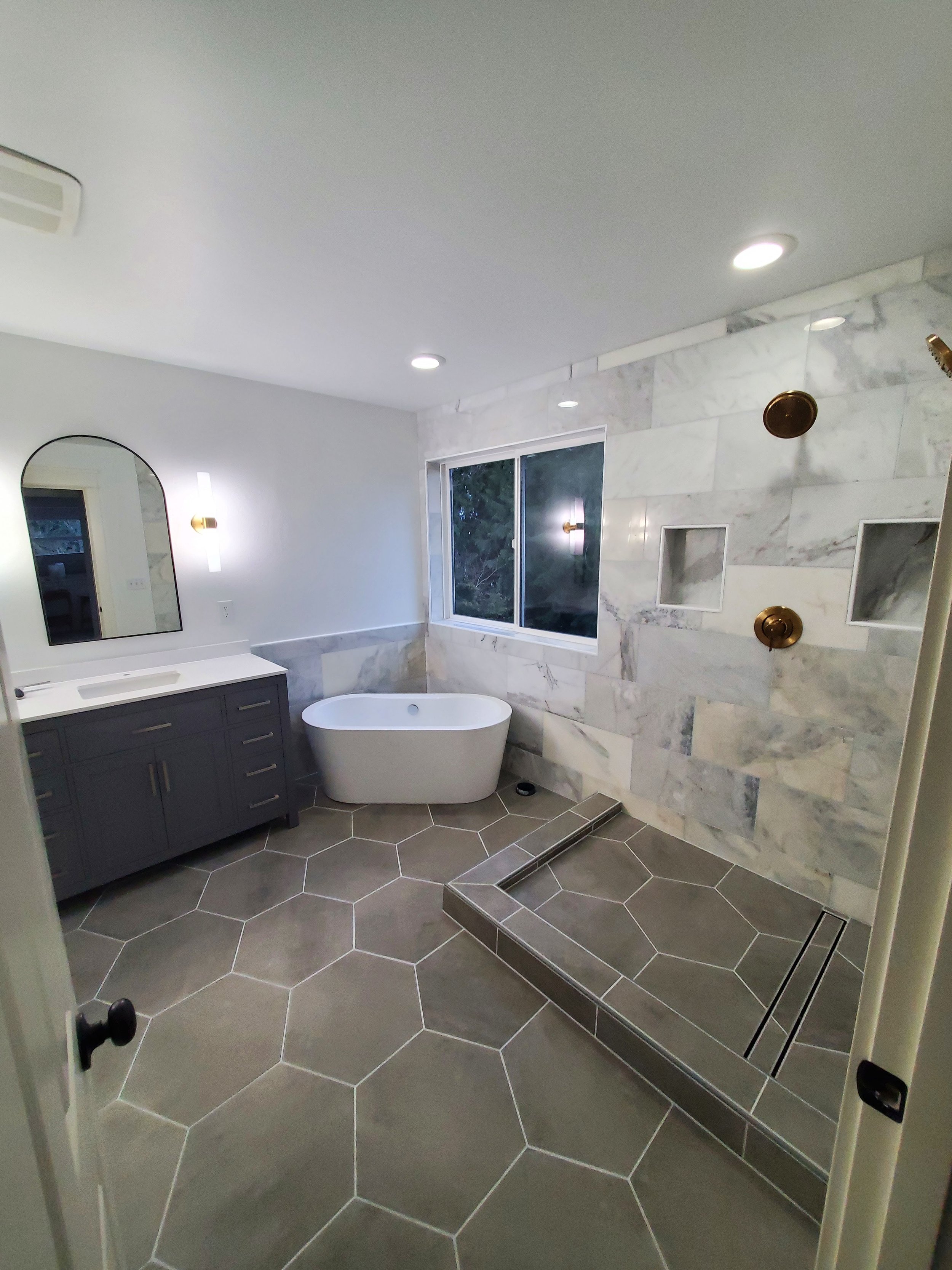 Modern bathroom with gray vanity, white sink, standalone bathtub, window, gray tiled floor with hexagonal tiles, marble tiled wall with built-in niches, and a walk-in shower with a linear drain and gold fixtures.
