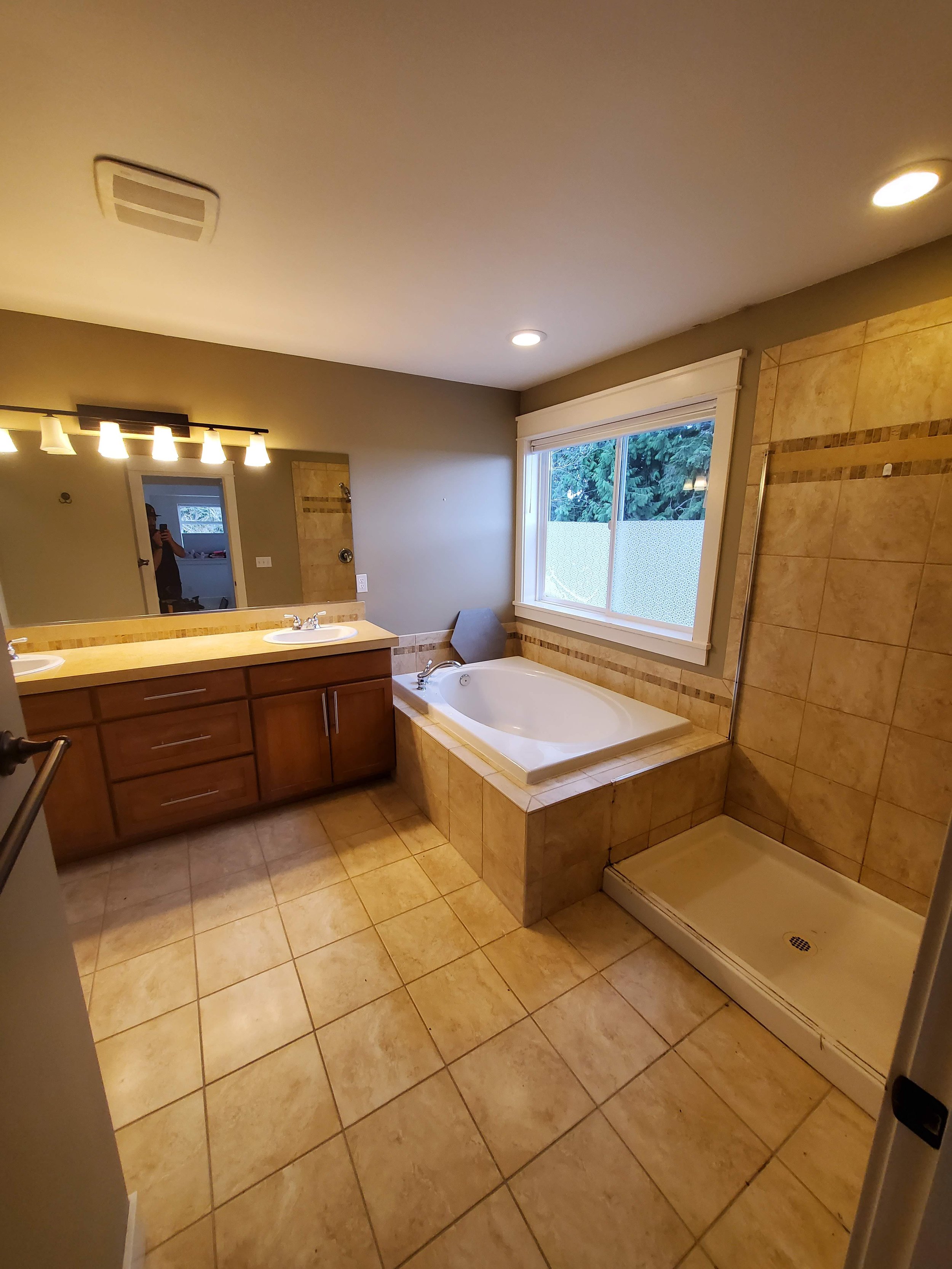 A bathroom with a double sink vanity, a large mirror, a bathtub, and a separate shower area with beige tiles and a window showing greenery outside.